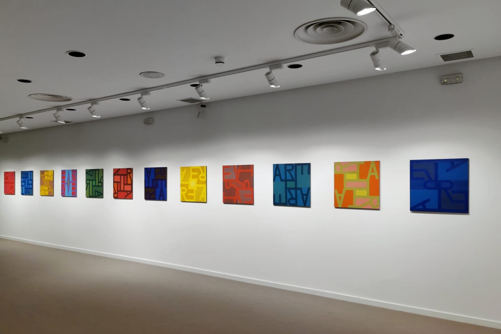 Serie de cuadros abstractos con letras de diferentes colores en una pared blanca de galería de arte.