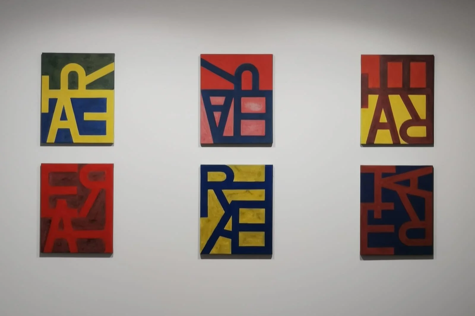Seis cuadros abstractos con letras y formas en colores rojo, amarillo, azul y verde, colocados en una pared blanca.