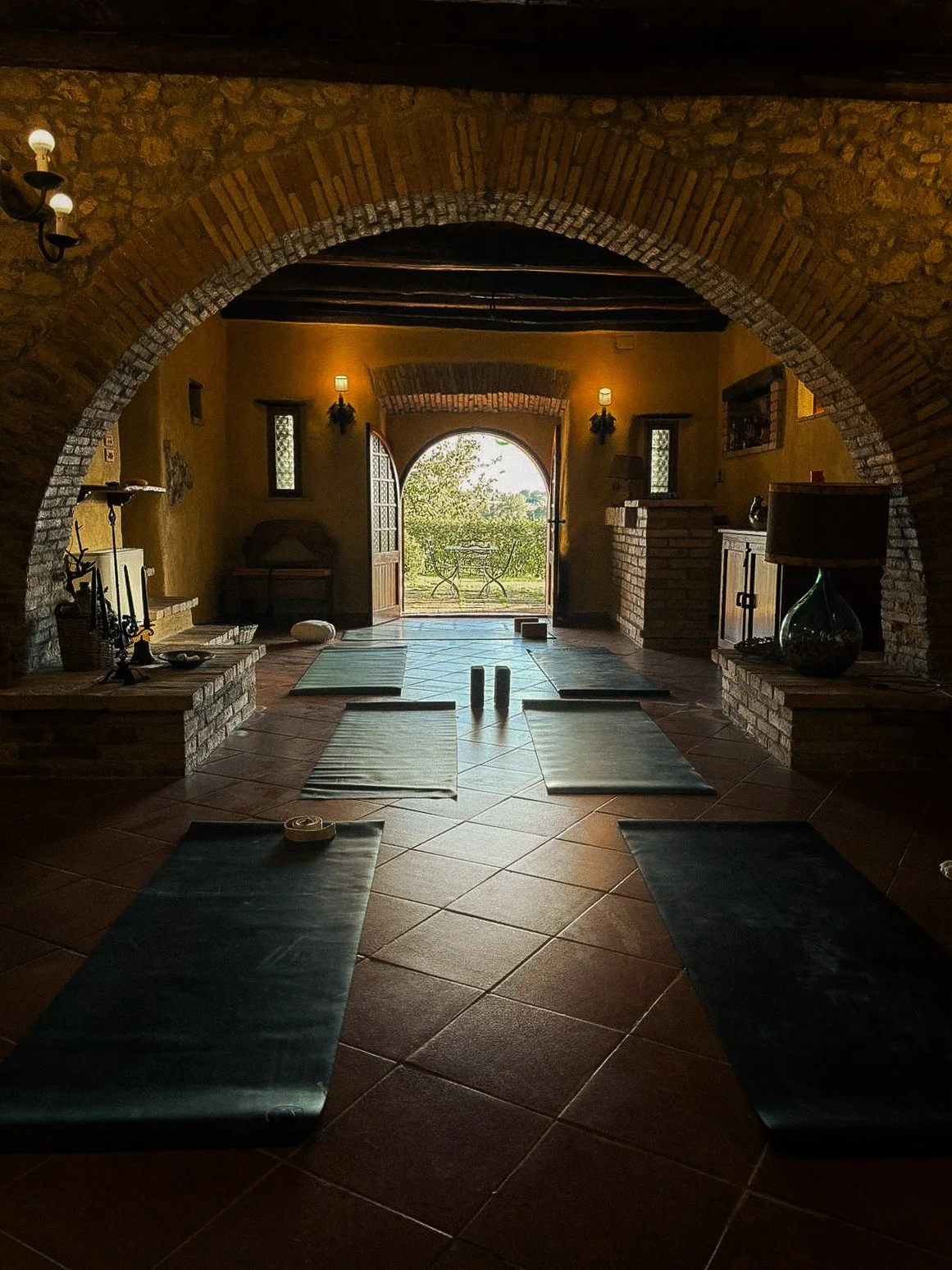 Yoga mats arranged on a tiled floor in a rustic room with a large archway opening to a balcony with outdoor furniture, warm yellow walls, and stone accents.