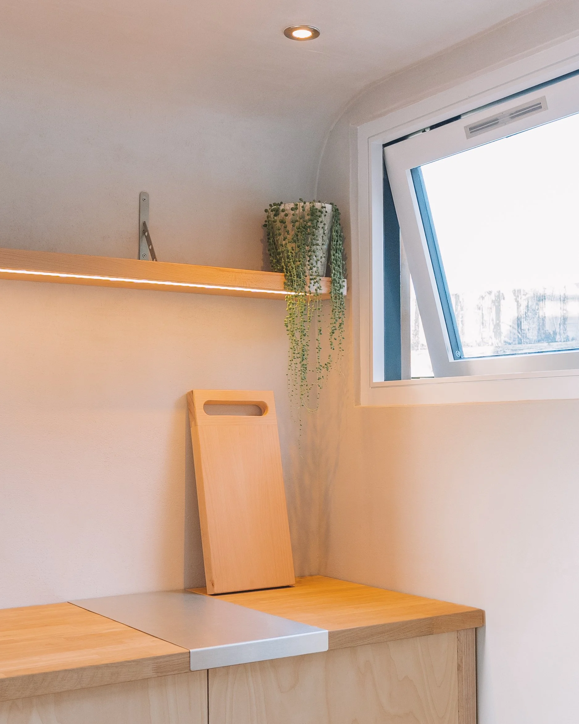A corner of a room with a wooden countertop, a wooden cutting board leaning against the wall, a small shelf with a potted trailing plant, and a window with an angled open pane.