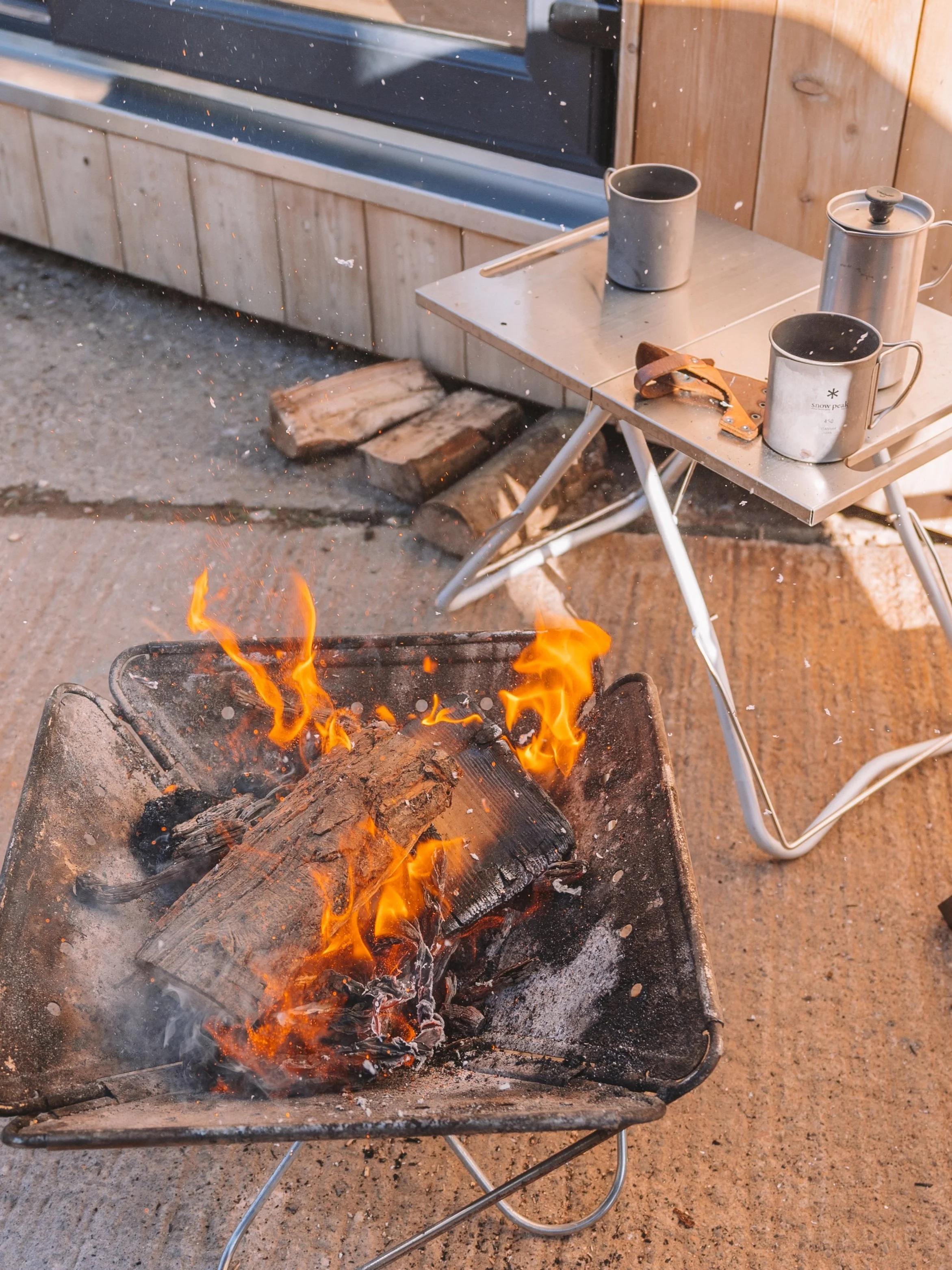 A small campfire with burning logs in a metal fire pit on a concrete surface next to a foldable table with camping mugs, a pot, and a leather strap, in a rustic outdoor setting.