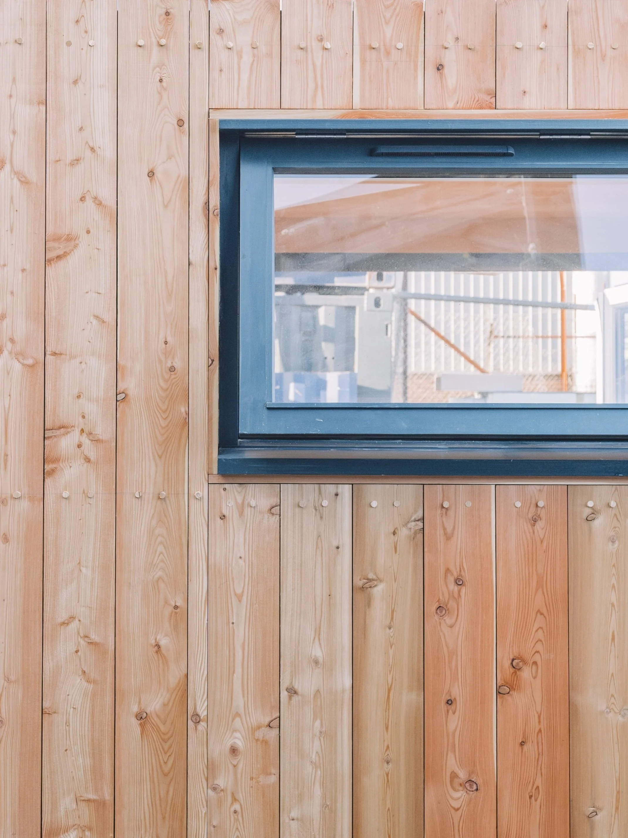 A window in a wooden wall with vertical planks and a dark frame, looking out onto a construction site with scaffolding and building materials.