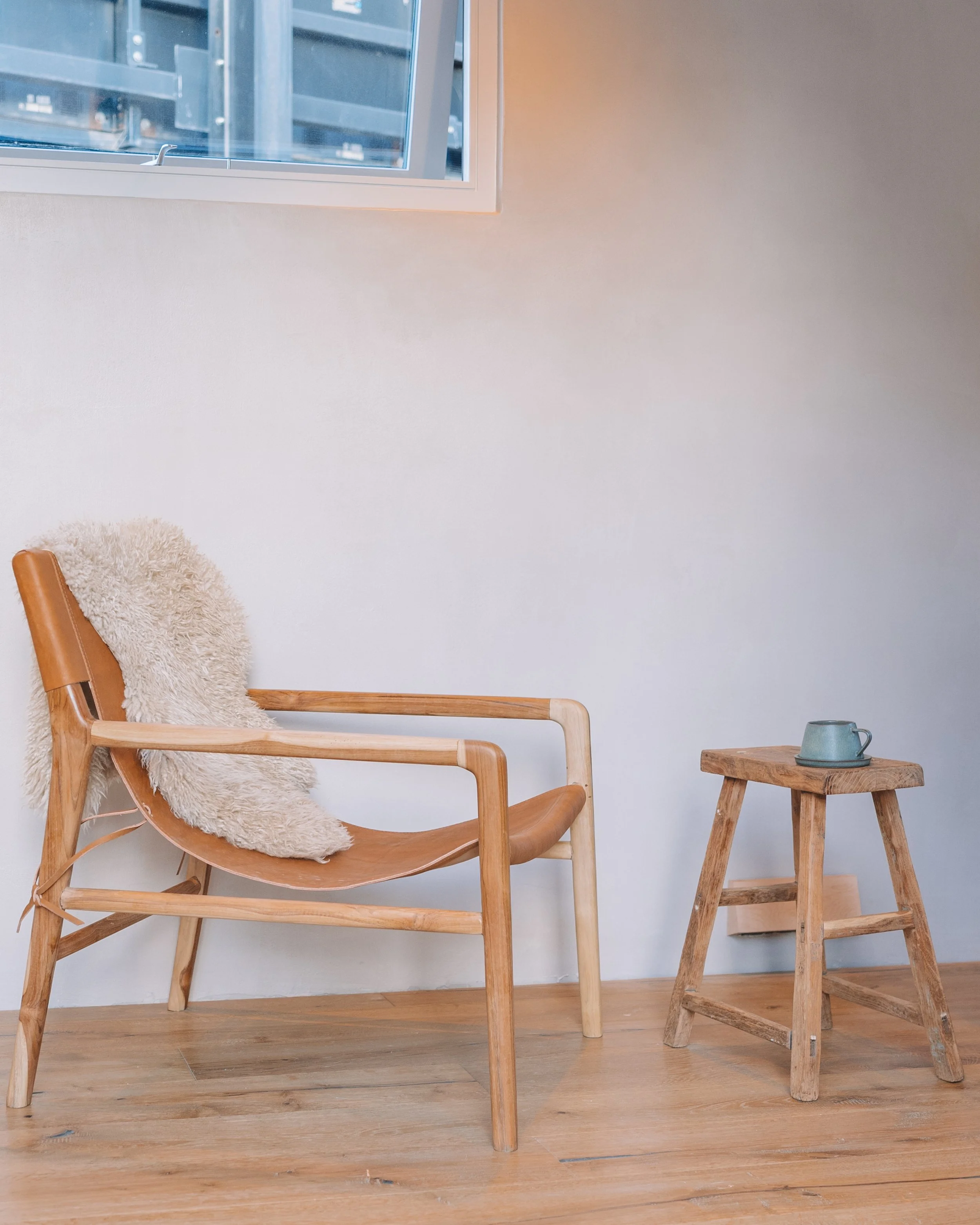 A wooden lounge chair with a sheepskin throw on it, next to a small wooden stool with a cup on top, against a plain white wall, with a window at the top left corner showing an urban exterior.