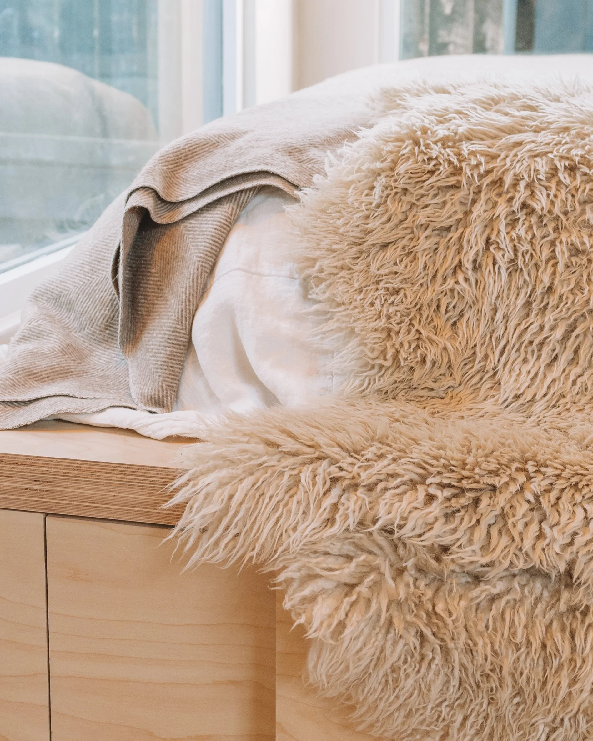Close-up of a cozy bed with a fluffy beige shaggy blanket, a white pillow, and a folded beige blanket, next to a large window with a view of the outdoors.