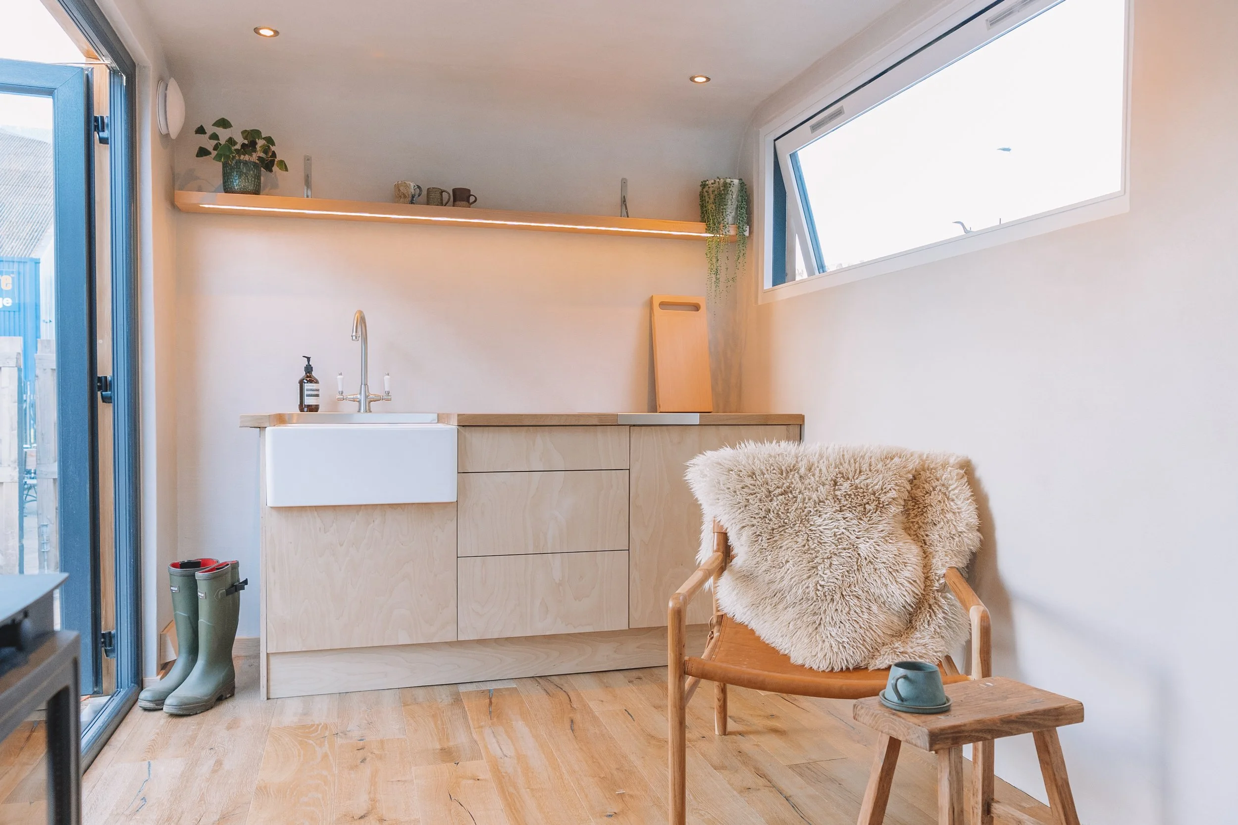 A cozy, minimalist kitchen area with light wood cabinetry, a white farmhouse sink, and a large angled window. There is a sheepskin throw draped over a wooden chair, which has a cup on a small wooden side table. Green boots are placed near the sink, a