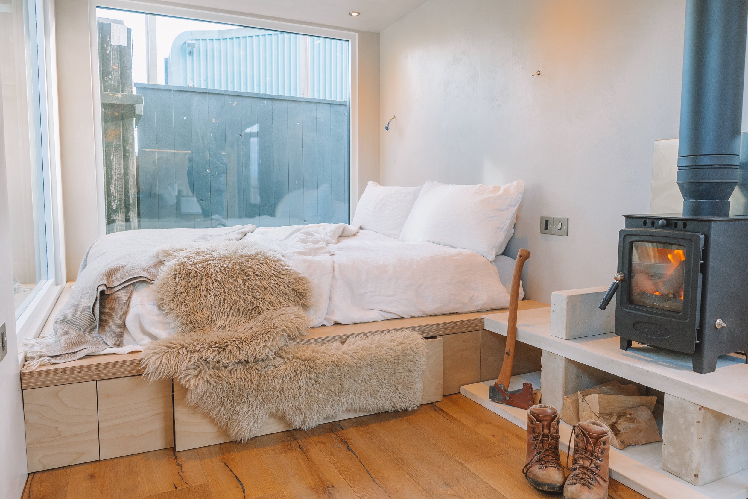 A cozy bedroom with a bed raised on a wooden platform, covered with white linens and a shearling blanket. There is a plush shearling rug on the floor and a large glass window behind the bed showing an outdoor space with a blue fence. To the right, a 