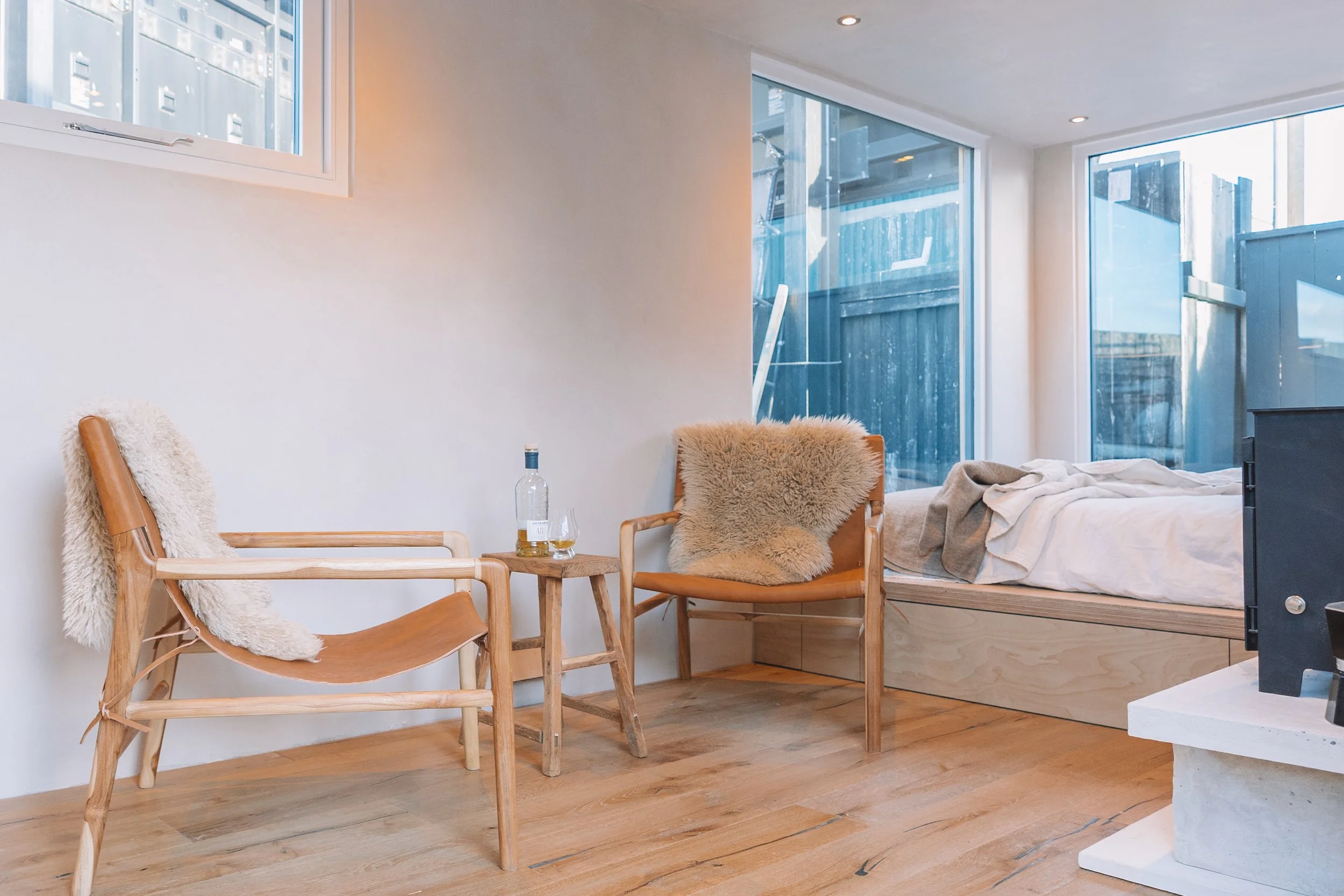 A cozy minimalistic bedroom with a bed near large windows, two wooden chairs with sheepskin throws, a small wooden side table with a bottle of alcohol and a glass, and a modern fireplace.