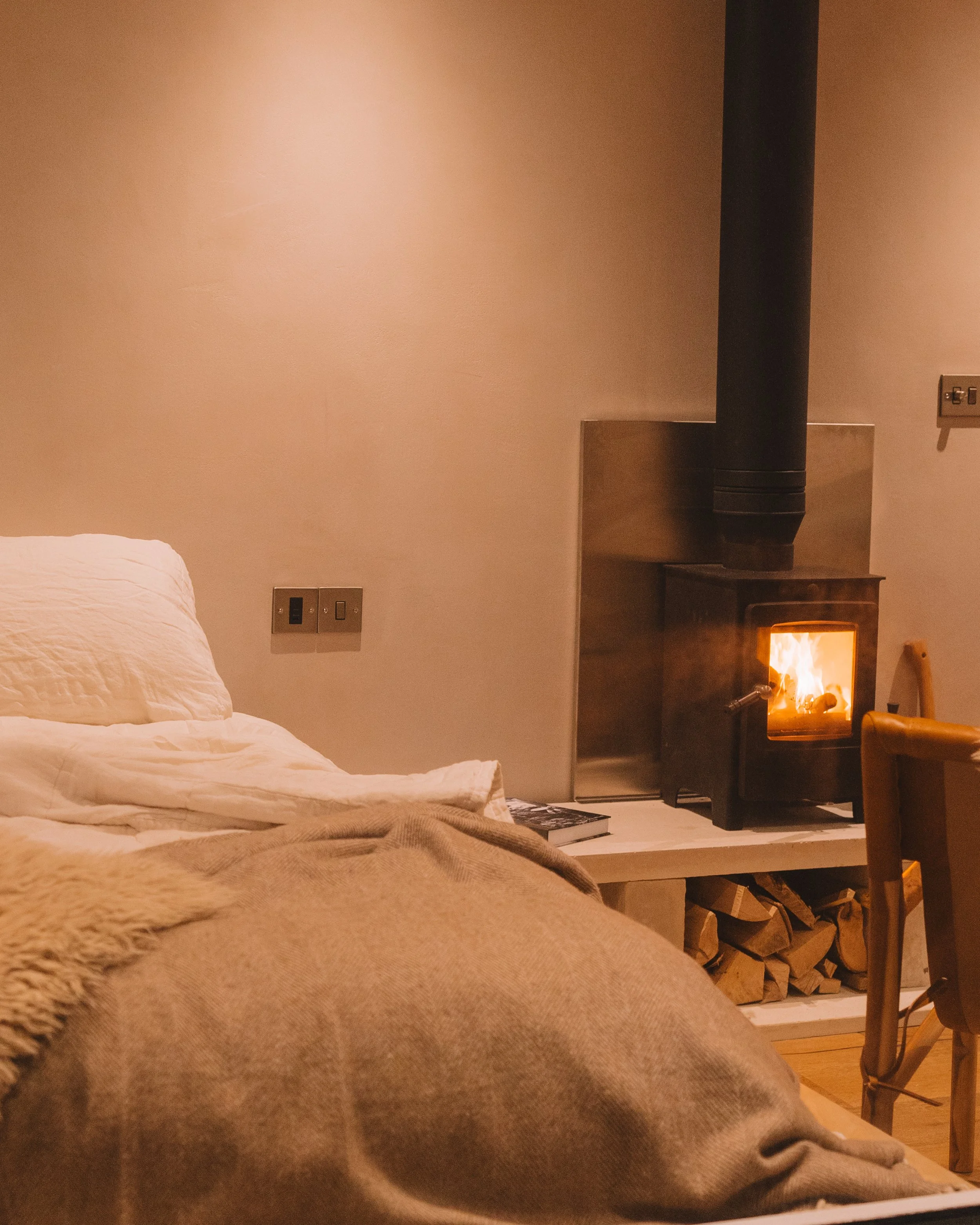 A cozy bedroom with a bed covered in beige blankets, a small wood-burning stove with a glowing fire, and a stack of firewood underneath a white shelf.