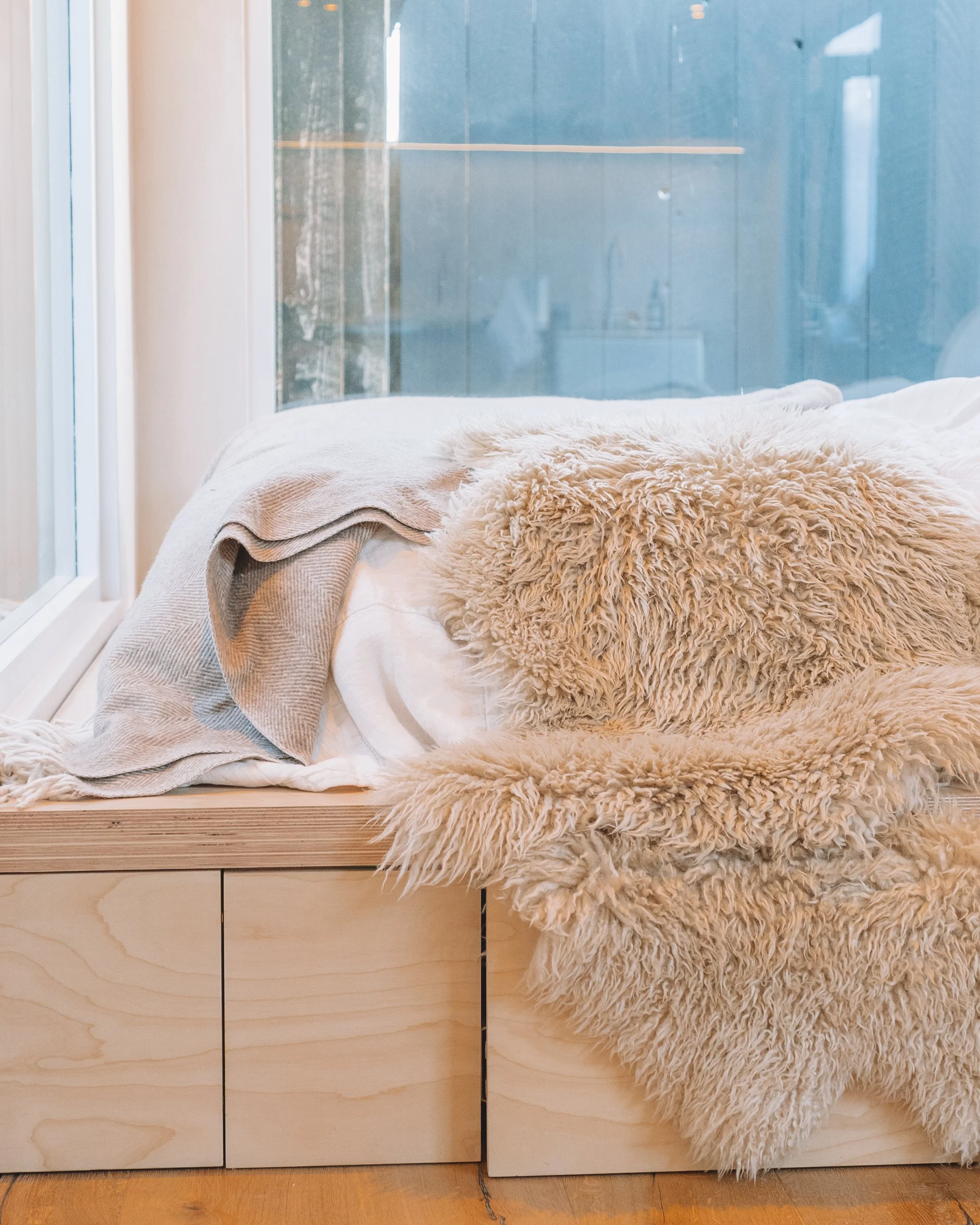 A cozy wooden window seat with a beige blanket, a textured beige pillow, and a sheepskin rug. There is a large window with a view of a modern building outside.