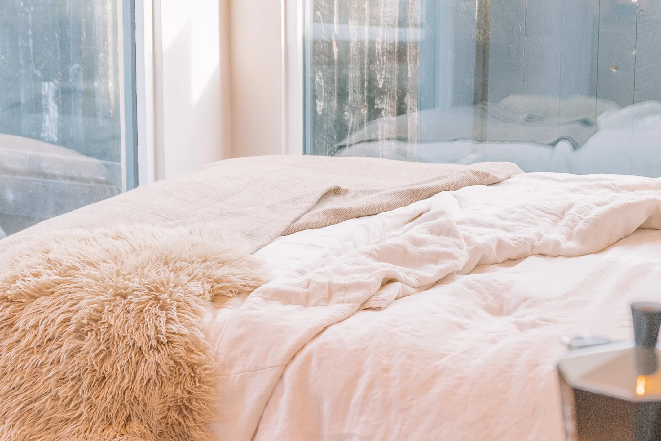 A cozy bed with white and beige linens, a fluffy beige throw blanket, and a glass wall in the background letting in natural light.