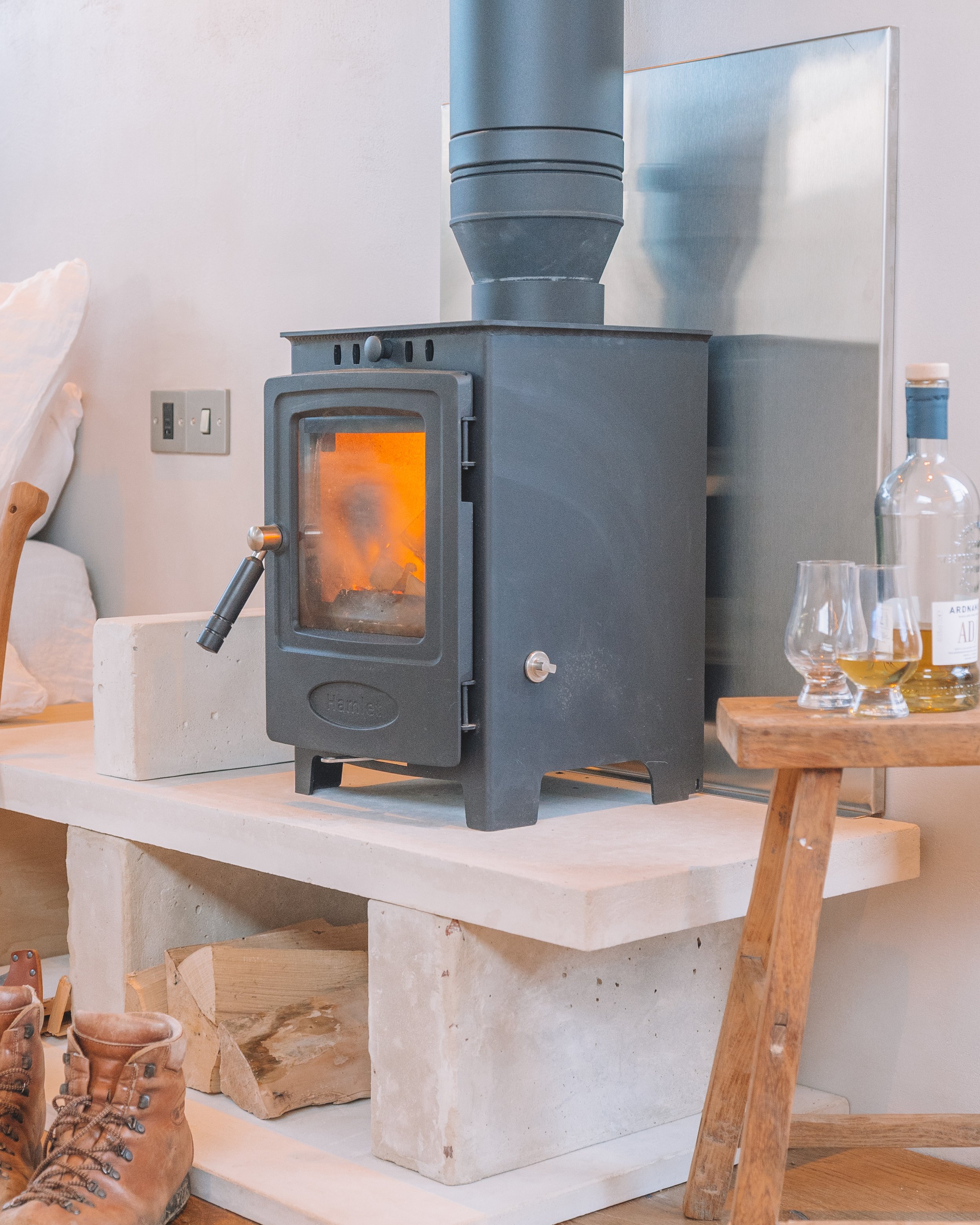 A black wood-burning stove with a glass door, on a concrete and wooden platform, with a pipe connected to the ceiling. A bottle of liquor and two glasses are on a small wooden table beside it.