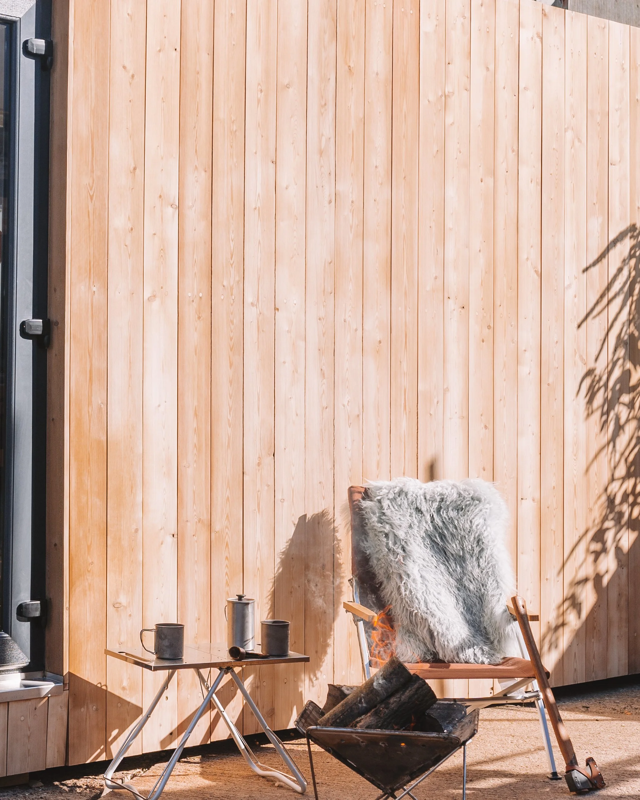 An outdoor scene with a wooden panel wall, a folding chair with a gray furry cushion, a small table with cups and thermoses, and a fire pit with logs.