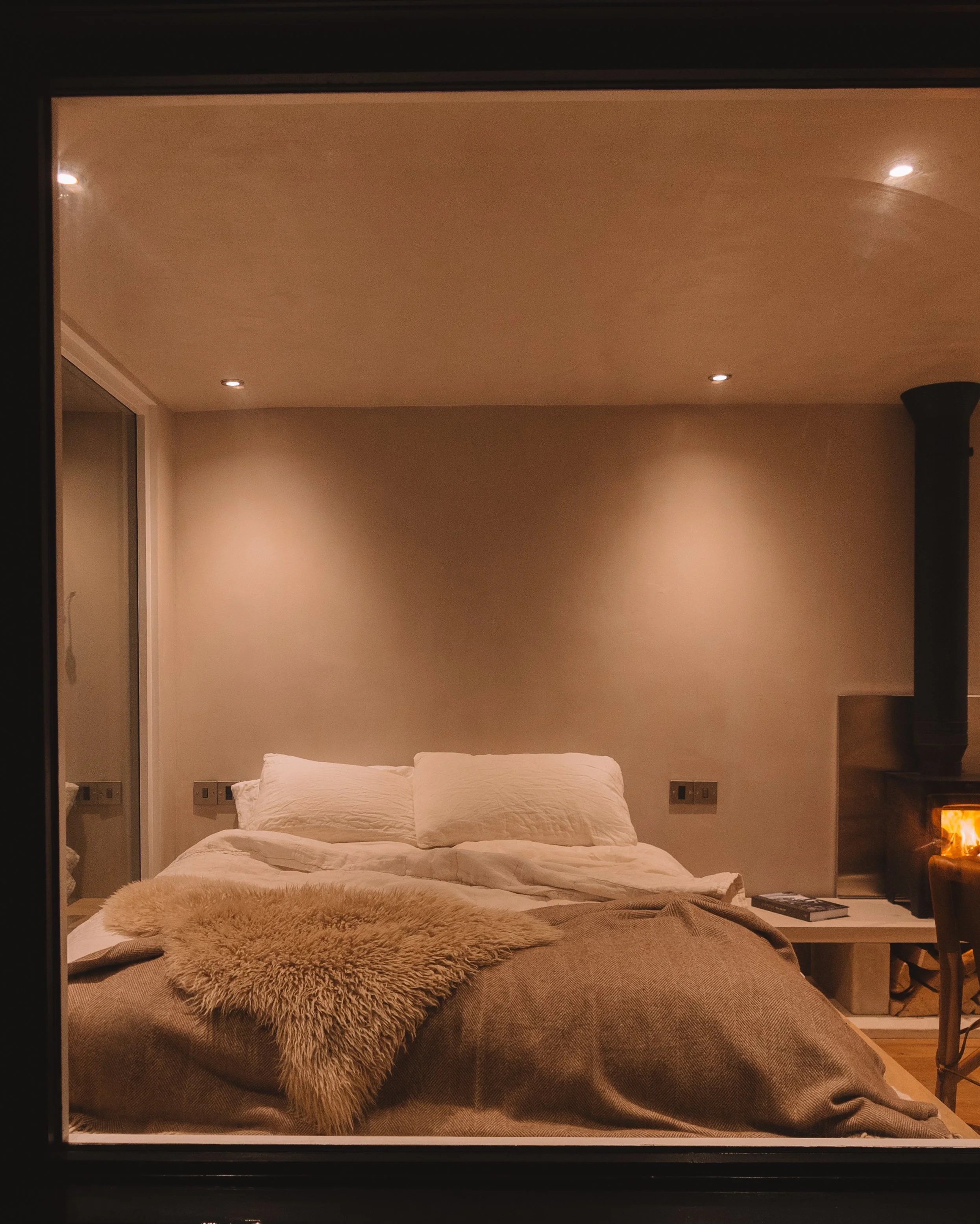 A bedroom with a bed, pillows, a furry blanket, a fireplace, and a chair, seen through a window or mirror.
