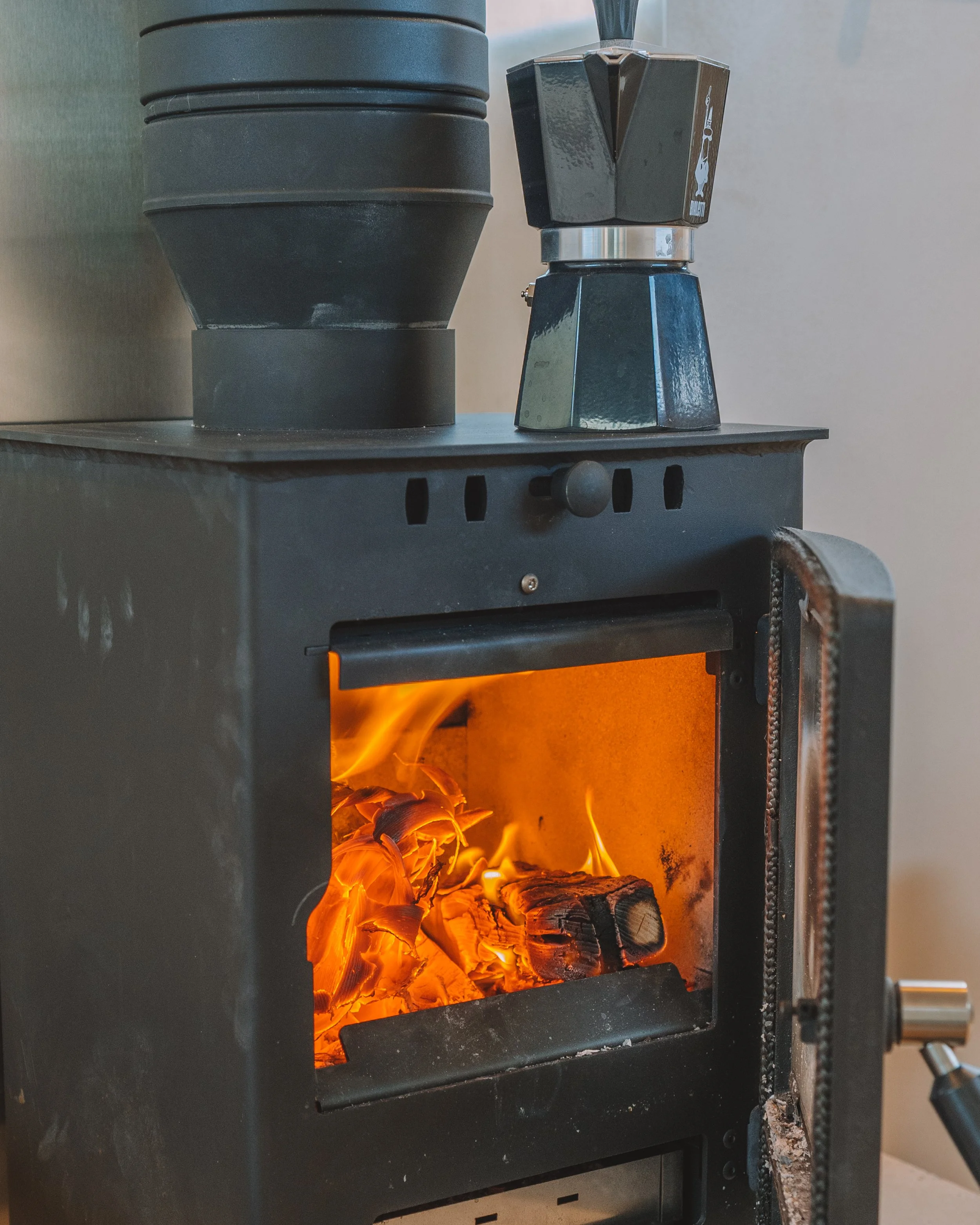 A small black wood stove with an open door showing glowing wood logs burning inside, and a moka pot placed on top of the stove.