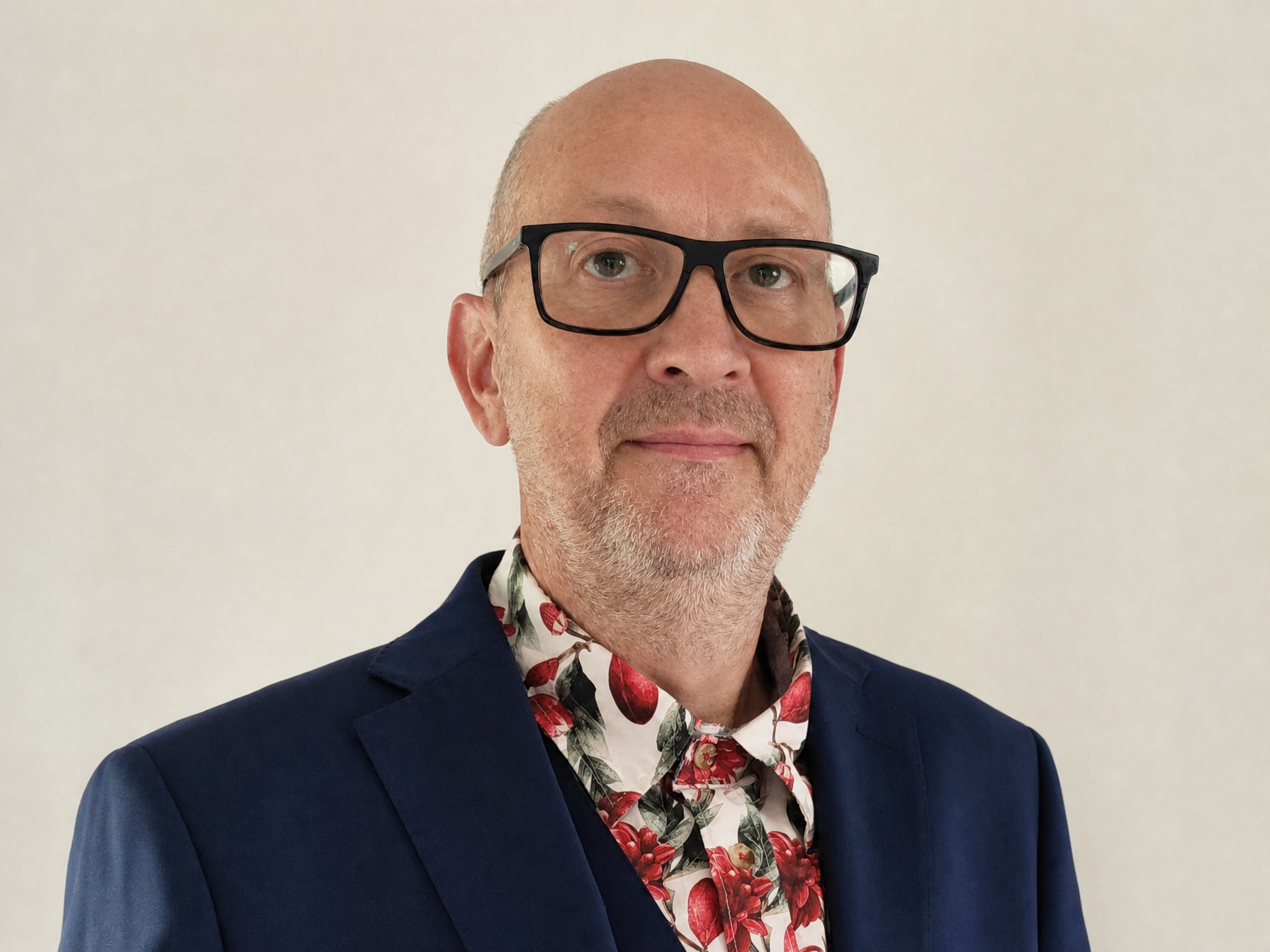 A middle-aged man with glasses and a short beard wearing a dark blue blazer and a floral shirt with red cherries stands against a plain light-colored background.