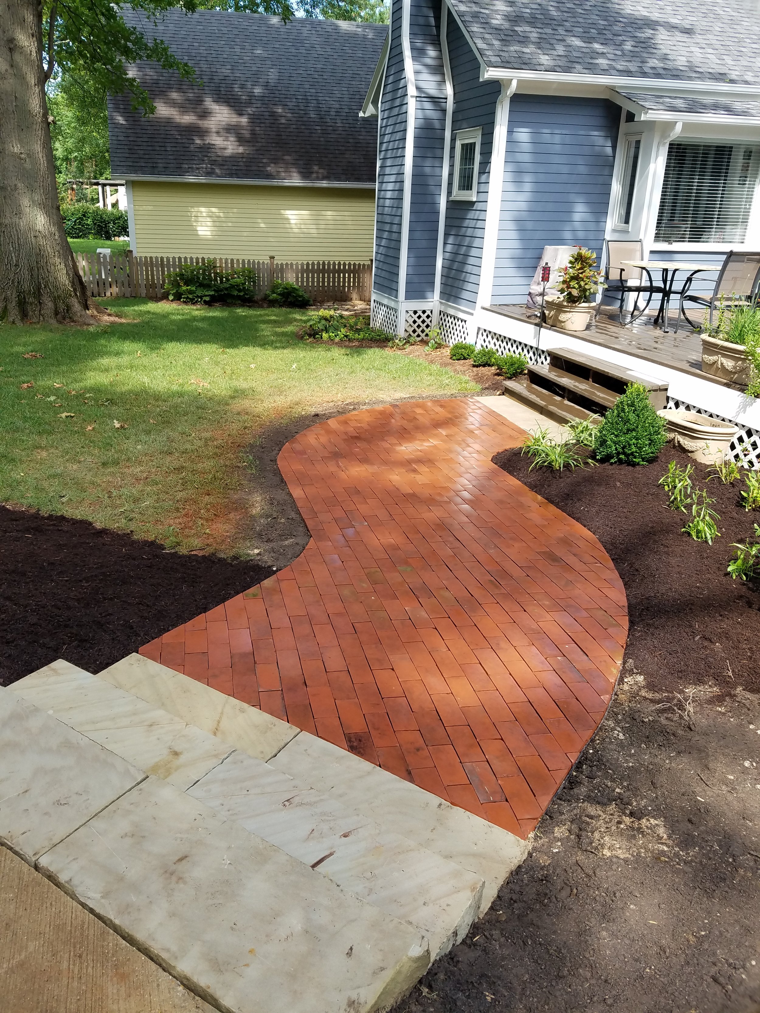 Curved red brick pathway leading to a blue house with a small porch, surrounded by a garden and lawn.