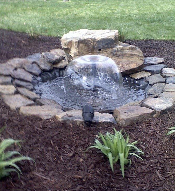 Small garden pond with rock border and bubbling water feature surrounded by mulch and plants.