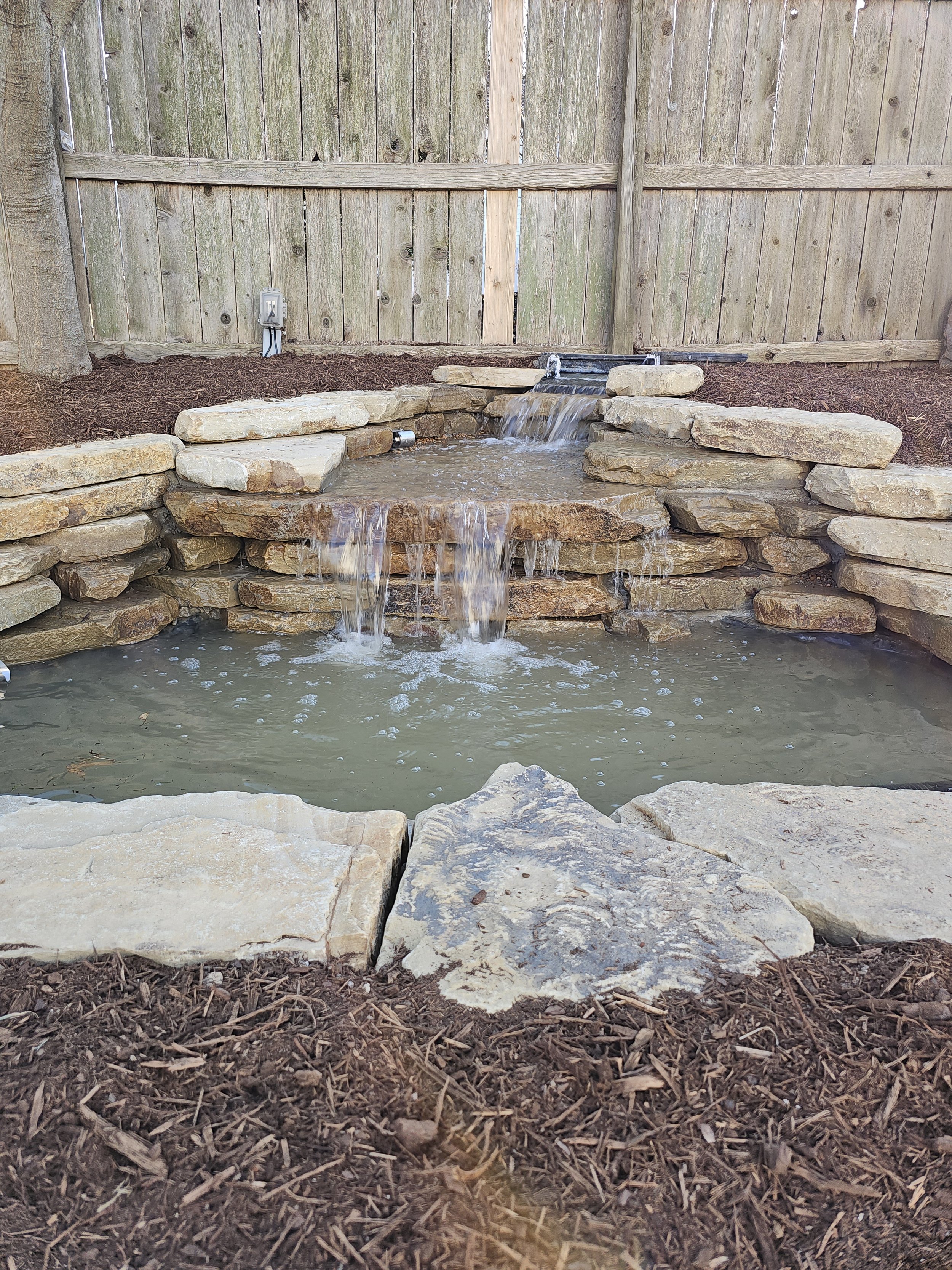 Small backyard waterfall with stone ledges and a wooden fence background.