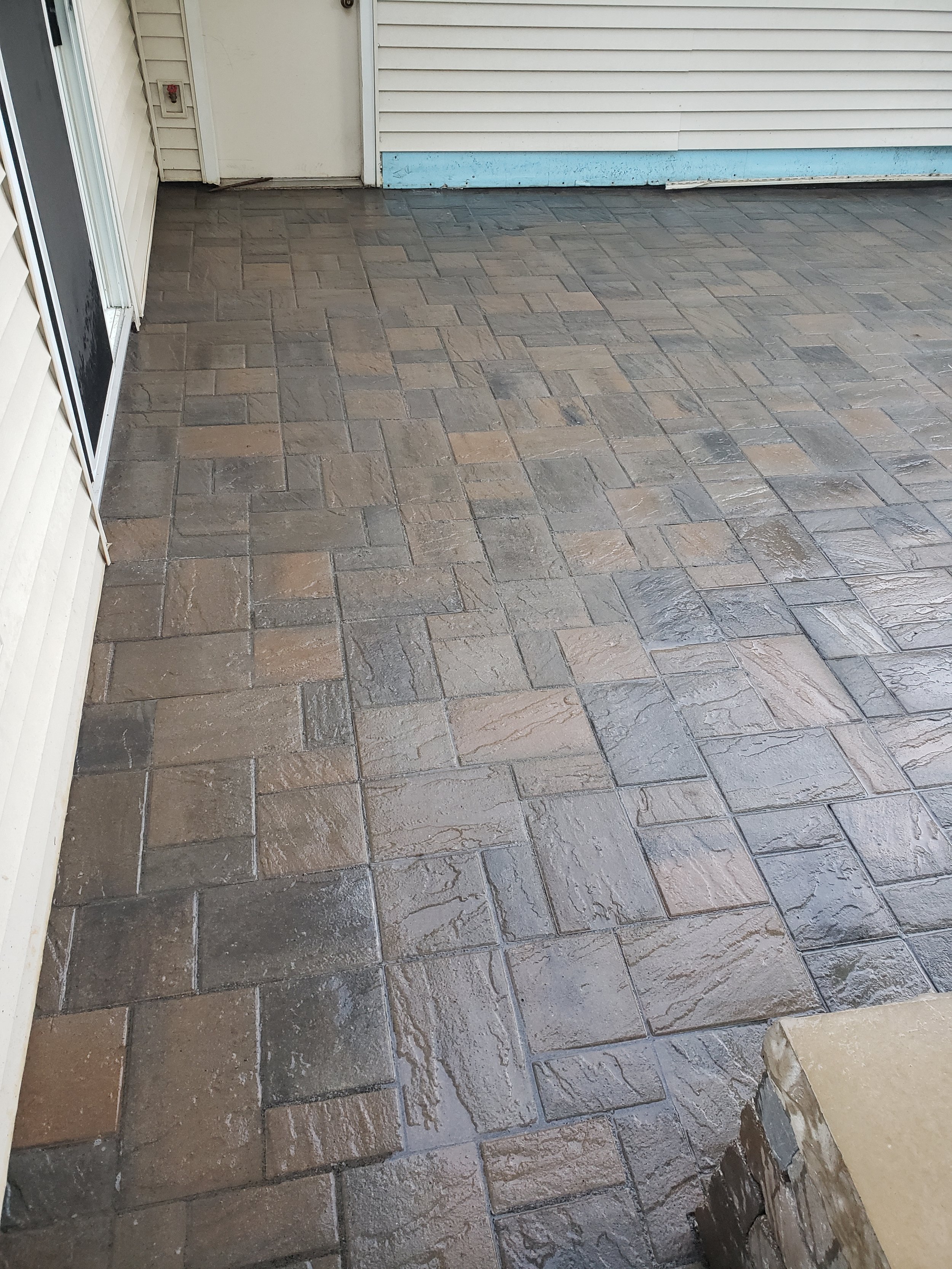 Patterned walkway with interlocking paving stones near a house with light siding and a door.