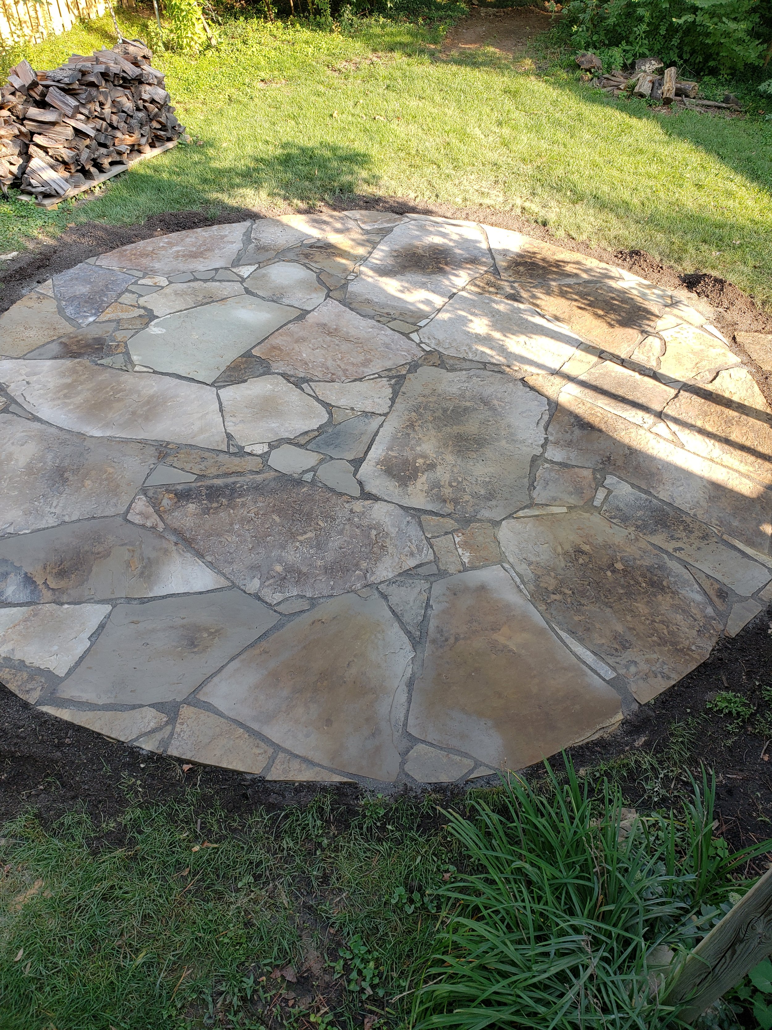 Circular stone patio in a grassy backyard with a stack of firewood nearby.