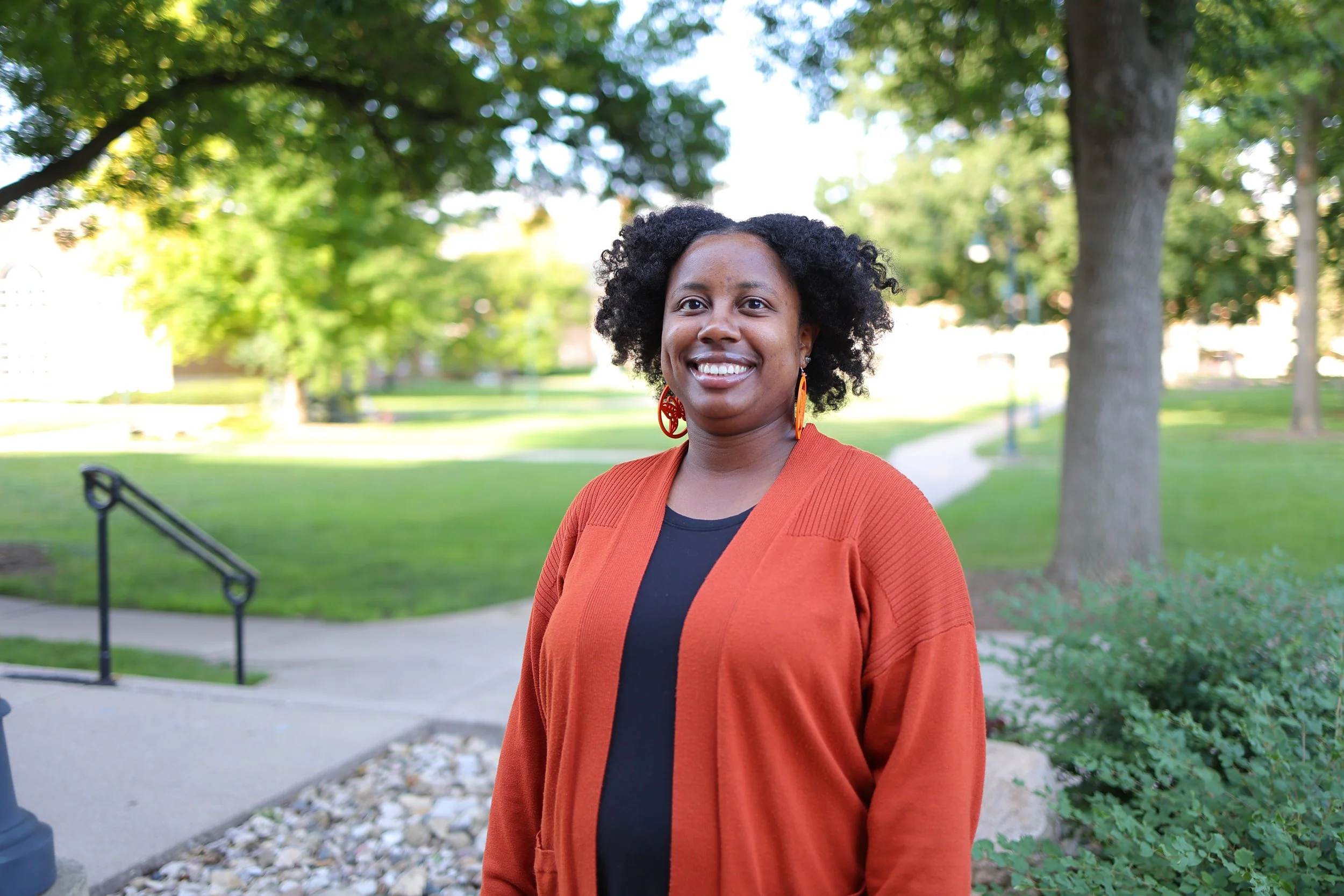 A woman with dark curly hair smiling outdoors, wearing an orange jacket over a black top, standing in a park with green trees and a pathway in the background.