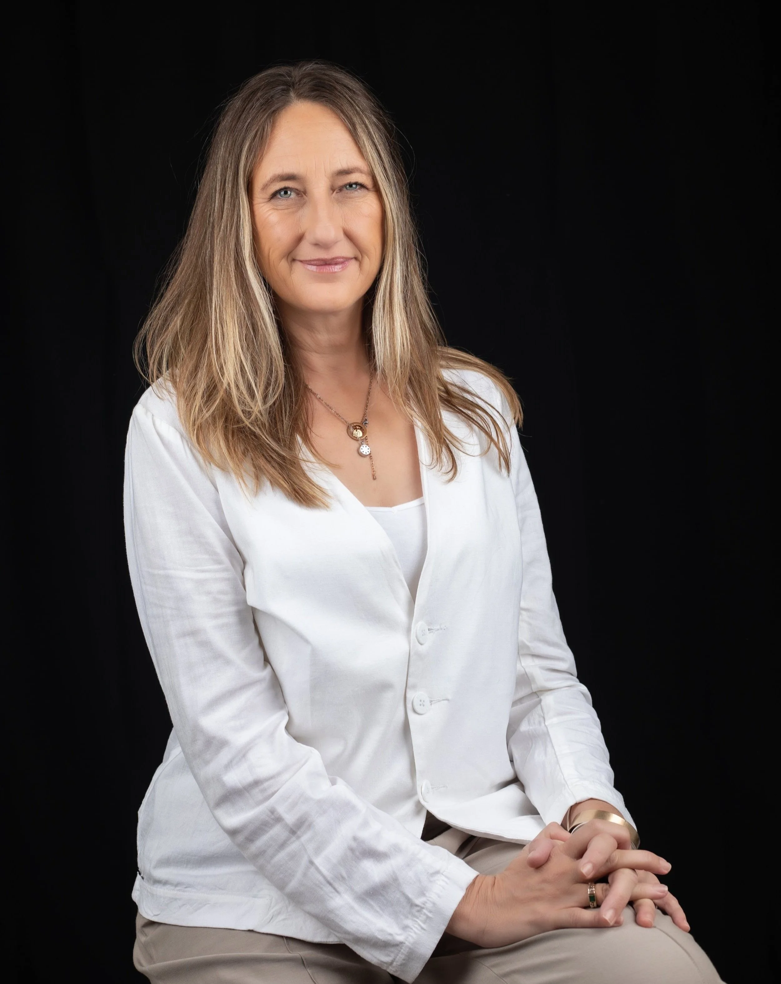 A woman with shoulder-length light brown hair wearing a white blazer, sitting against a black background, smiling softly. Salome van den Berg