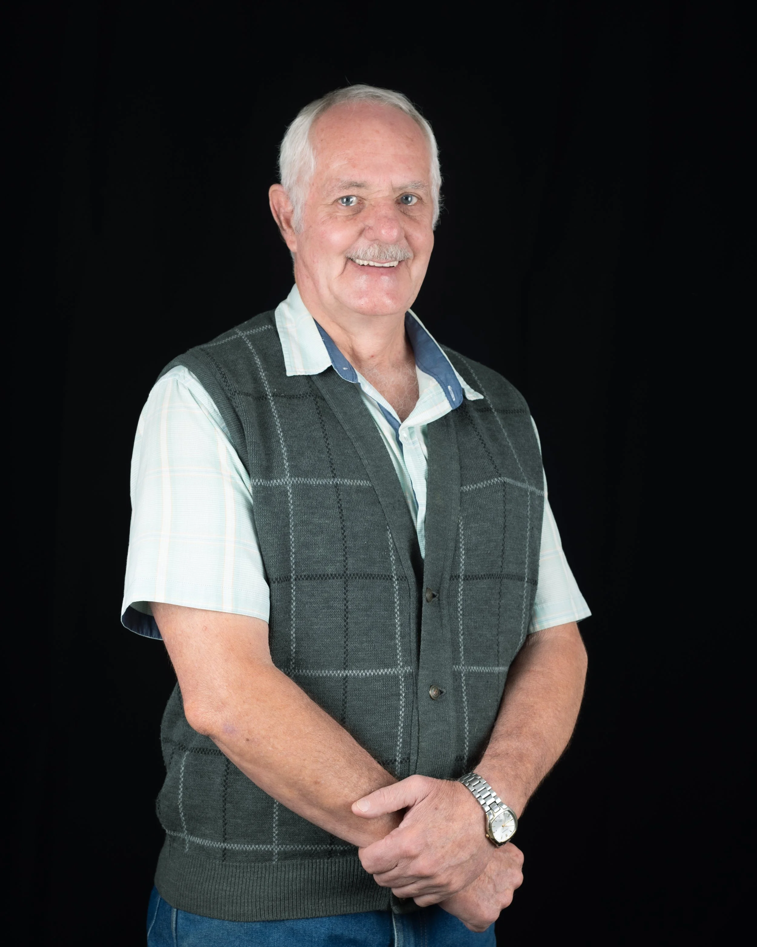 An elderly man with white hair and a mustache smiling, wearing a short-sleeved light blue shirt and a green plaid vest, standing against a black background. Dawie Steyn