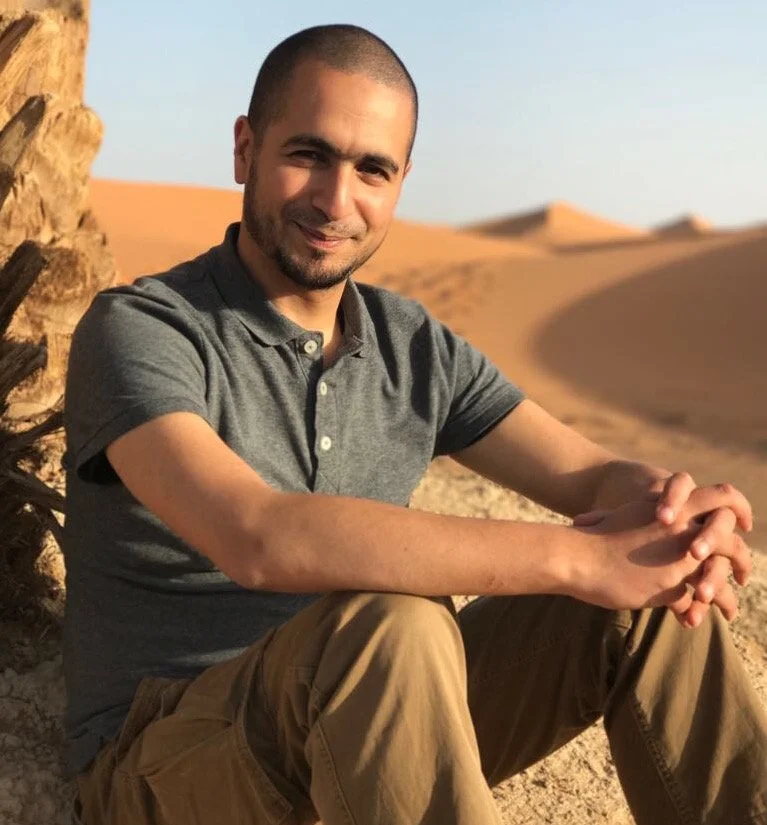 Nadir Moubarrid sitzt entspannt in einer Wüstenlandschaft, mit einem Palme im Vordergrund.