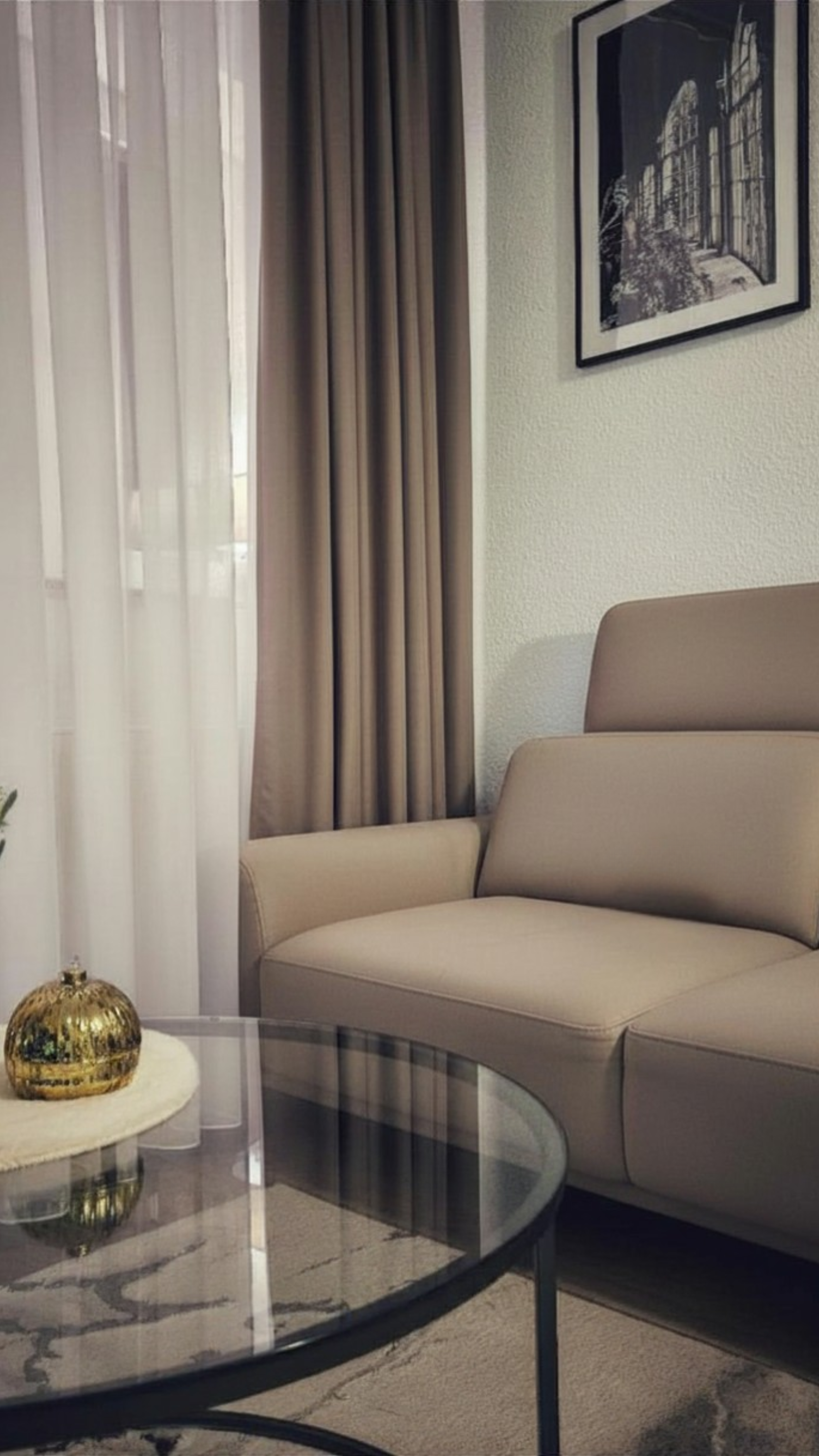 Ein Wohnzimmer mit einer beige Couch, einer Glasvitrine, einem Marmortisch mit einer goldenen Dekoration, gefasst von weißen Vorhängen, einer Wand mit einem schwarzen und weißen Stadtbild und einer Kachel-Vorhang.