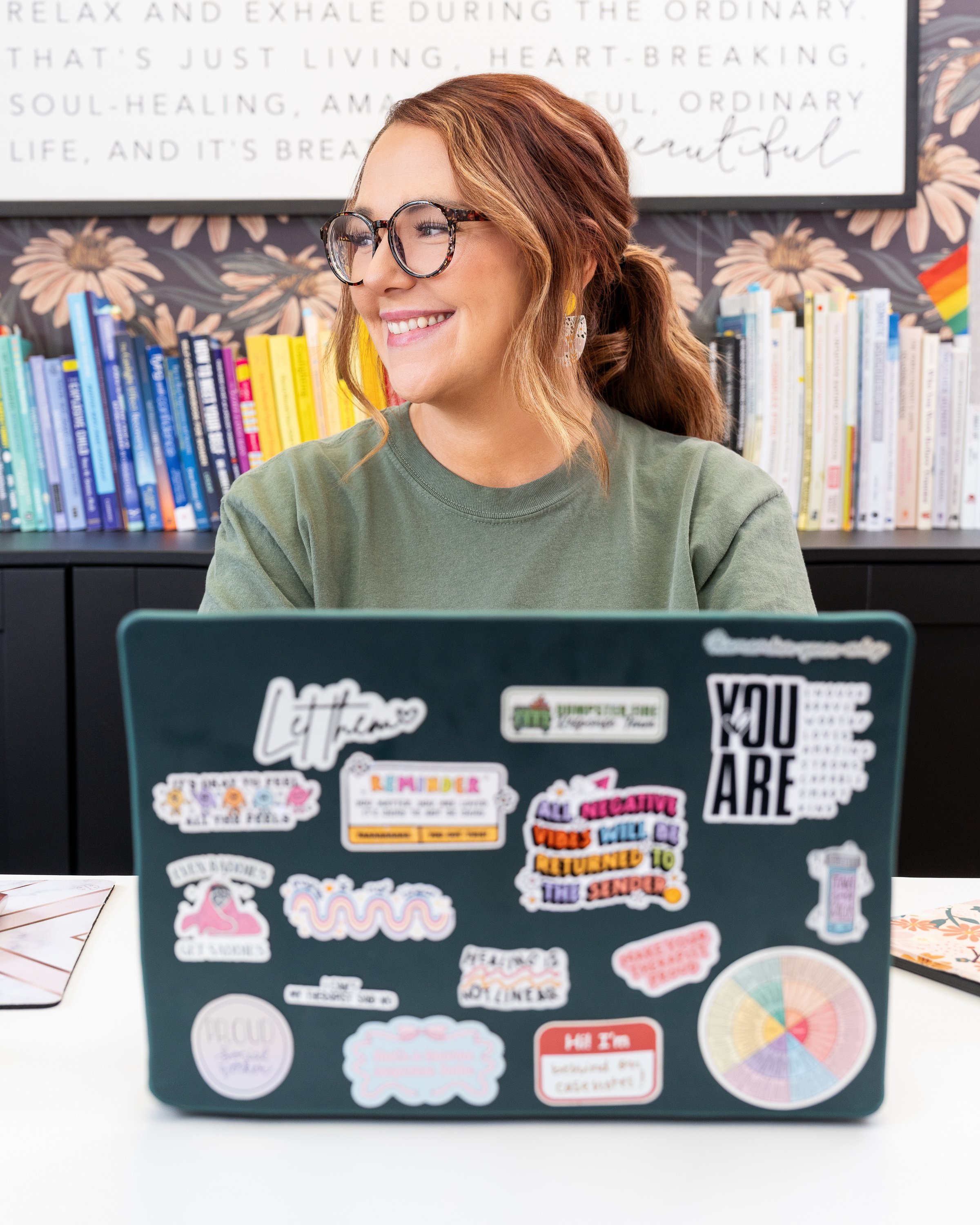 A smiling woman with glasses and earrings, sitting at a desk with a laptop covered in colorful stickers, in front of a bookshelf filled with books and a decorative wall.