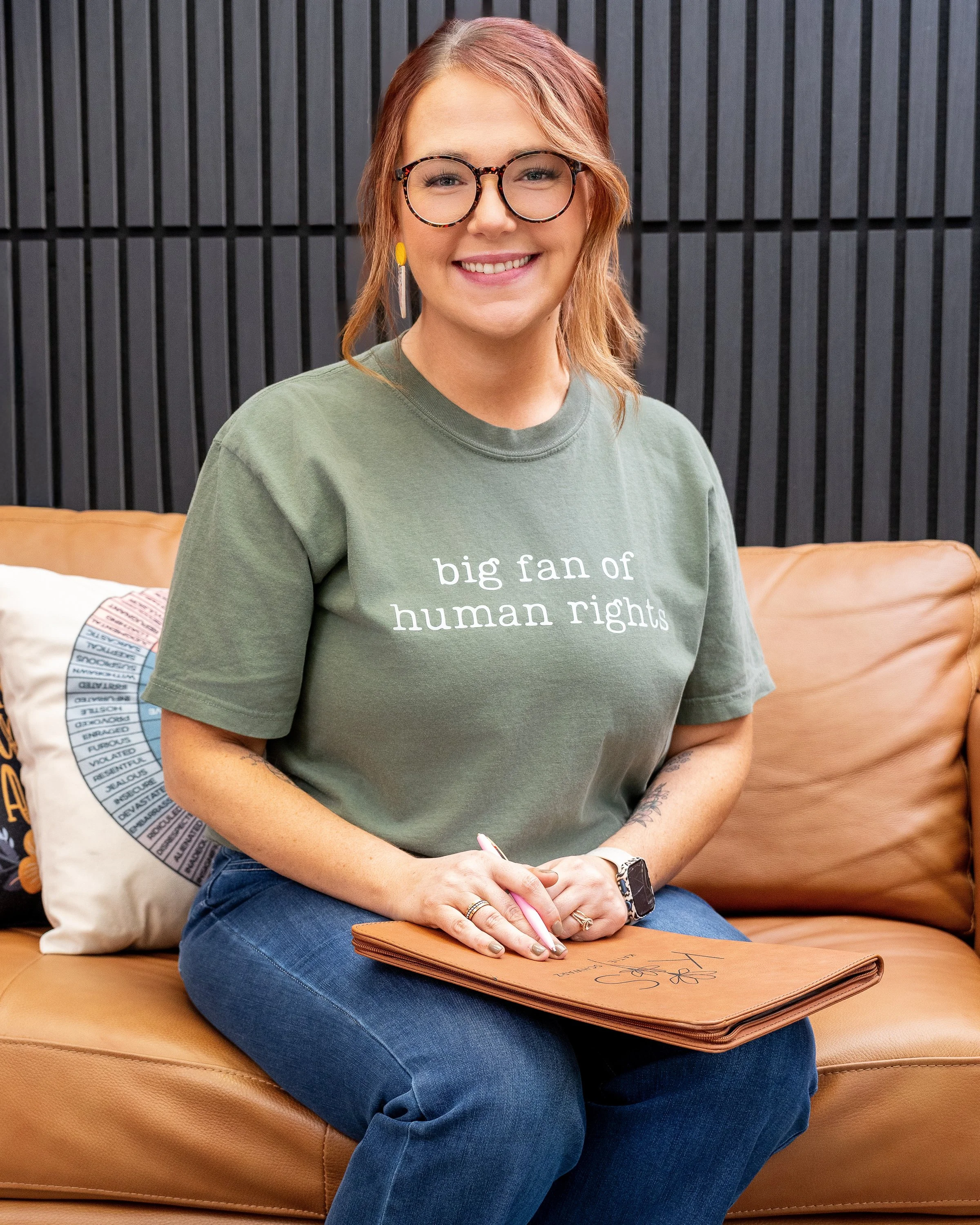 A woman with glasses and reddish hair sitting on a tan leather couch, smiling, wearing a green t-shirt that says 'big fan of human rights,' with a notepad or folder on her lap, with a black background and a decorative pillow.