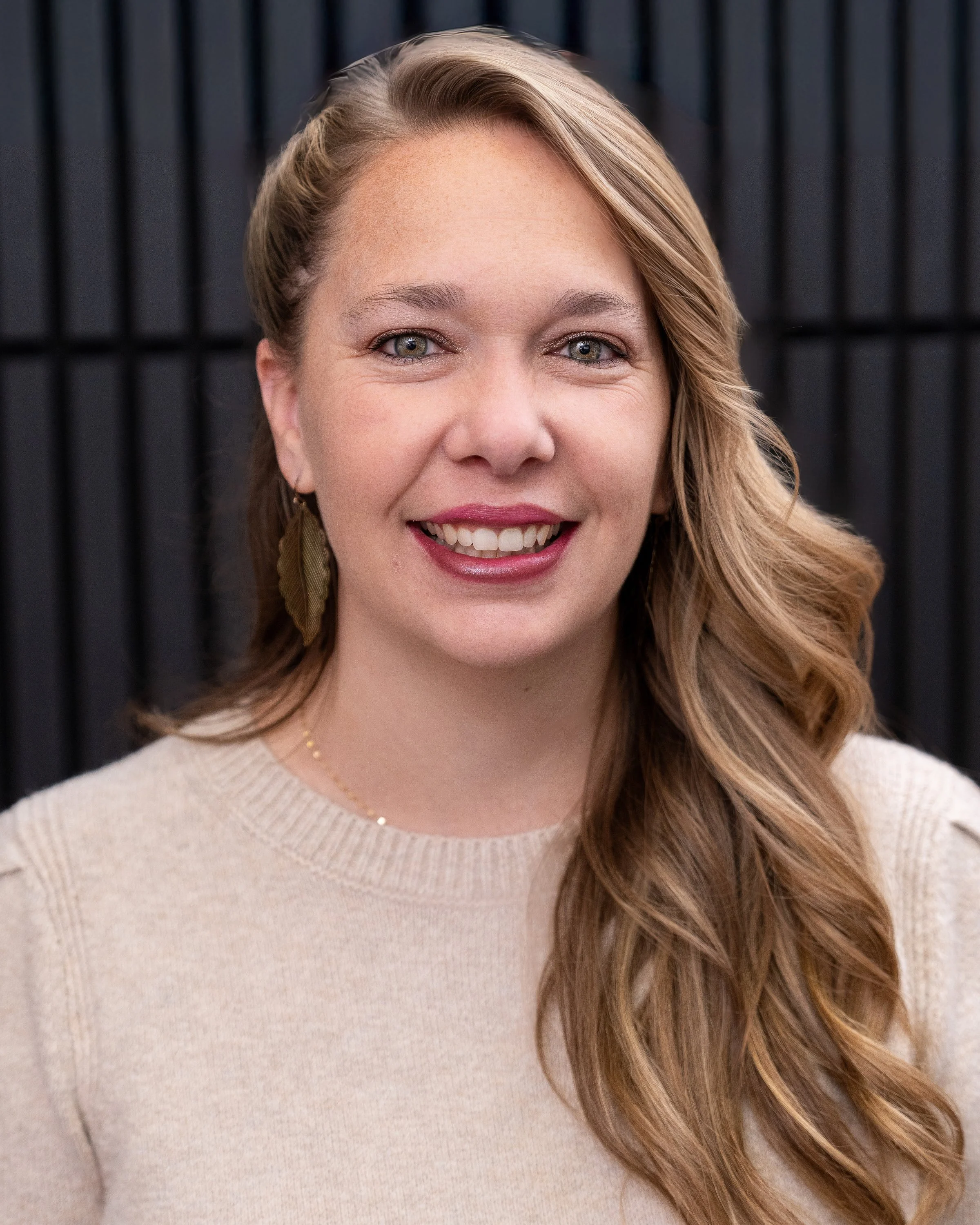 A woman with long, wavy, light brown hair, wearing a beige sweater, gold leaf earrings, and a gold necklace, smiling at the camera against a dark background.