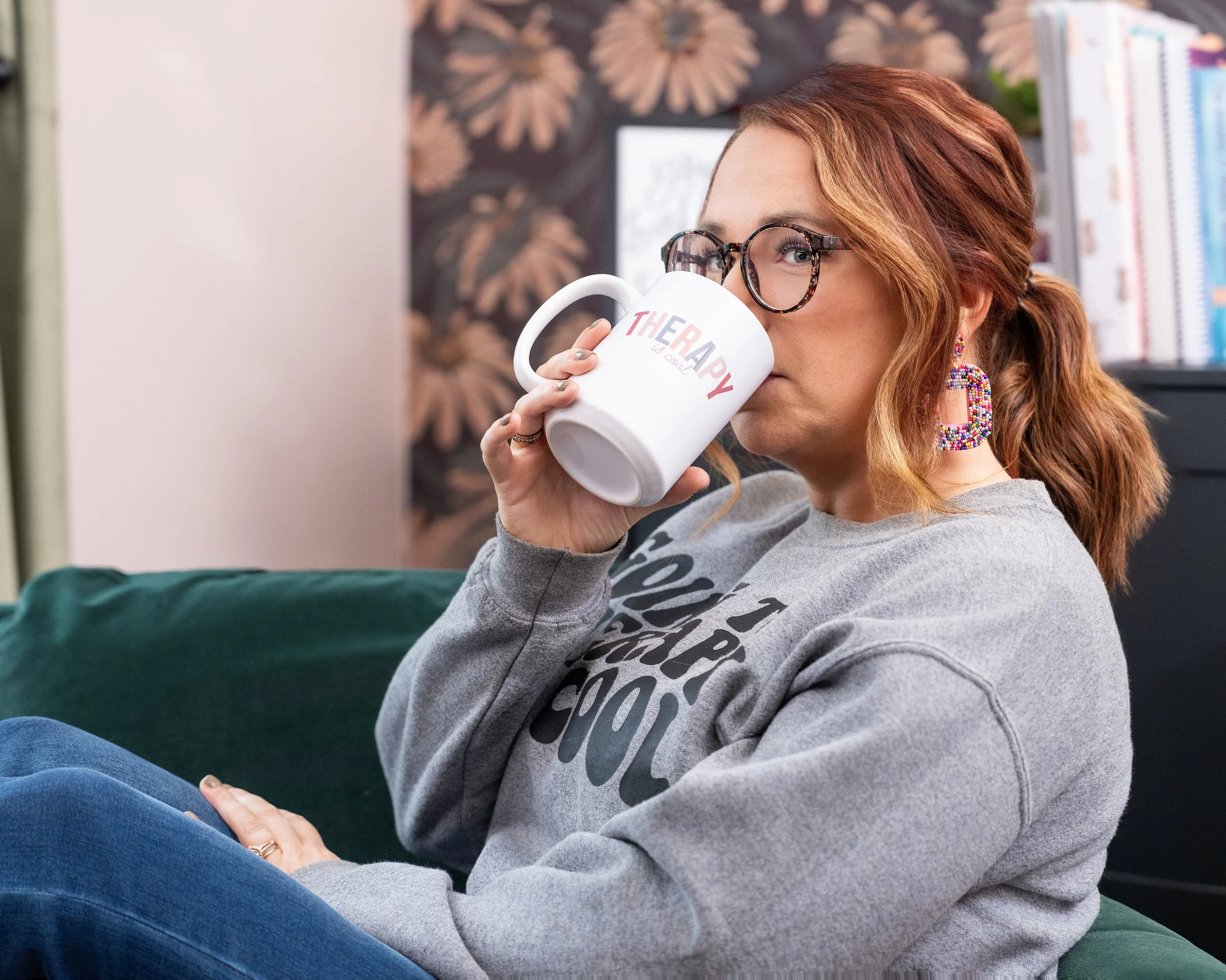 A woman with glasses and earrings is sitting on a couch, drinking from a white mug labeled 'THERAPY is cool'. She has red hair tied back, is wearing a gray sweatshirt and blue jeans, and is in a room with a floral wallpaper and bookshelves.