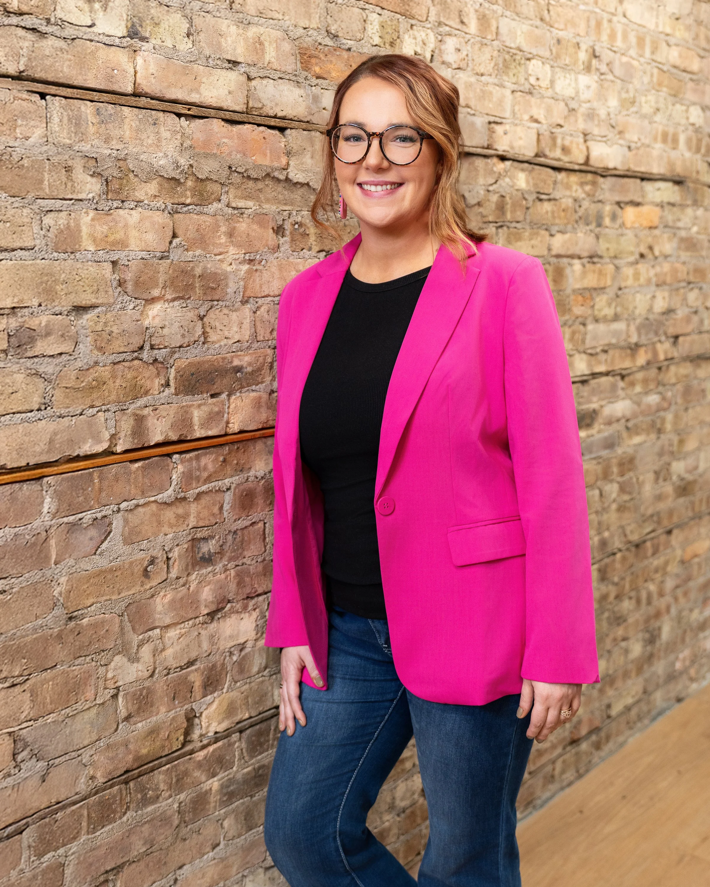 A woman with glasses and shoulder-length hair, wearing a bright pink blazer, black shirt, and jeans, standing against a brick wall and smiling.