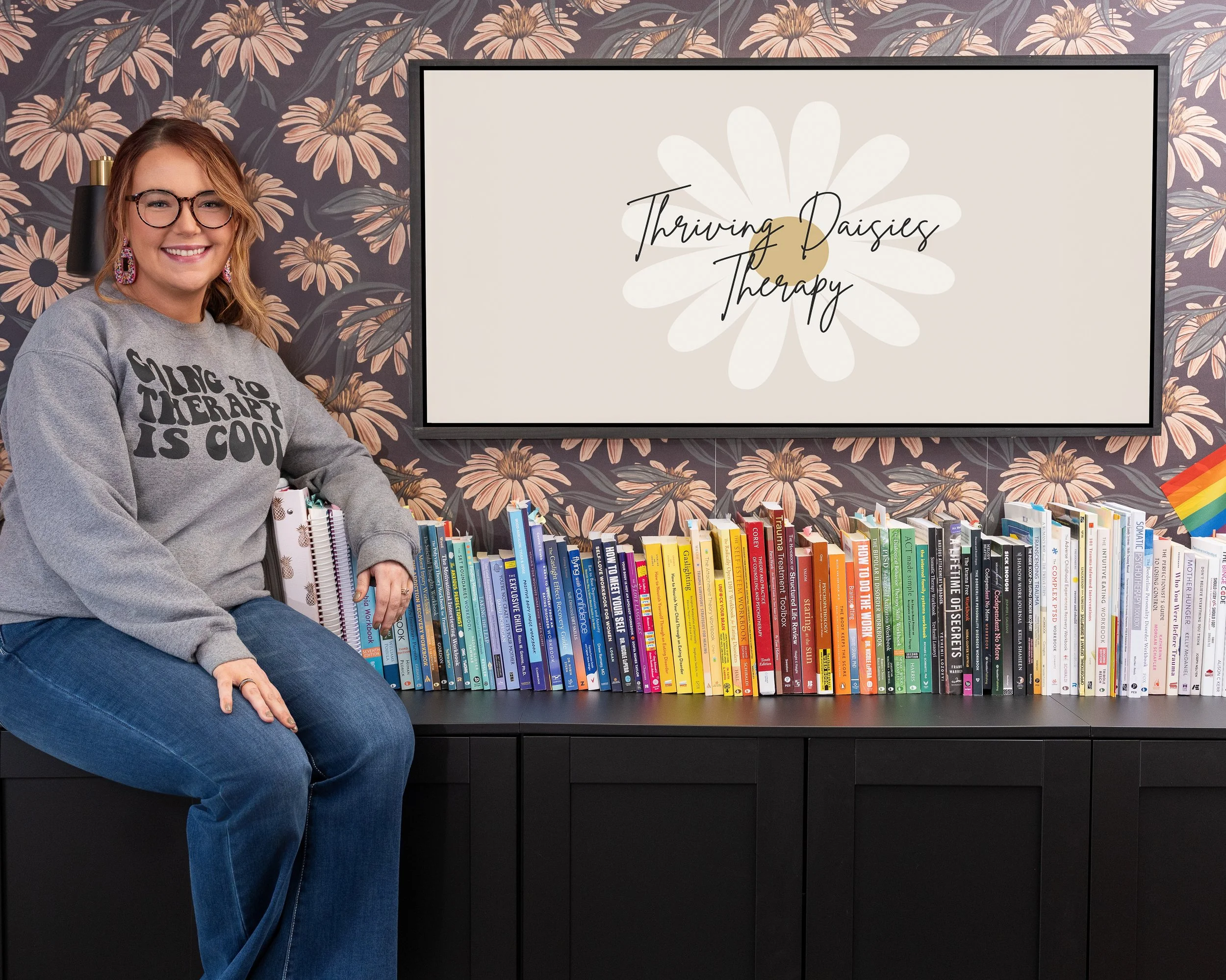 A woman with glasses and earrings, smiling and sitting on a black cabinet, is wearing a gray sweatshirt that says 'Singing to Therapy is Cool'. She is in a room with floral wallpaper and a large screen behind her that has a white daisy and the words 