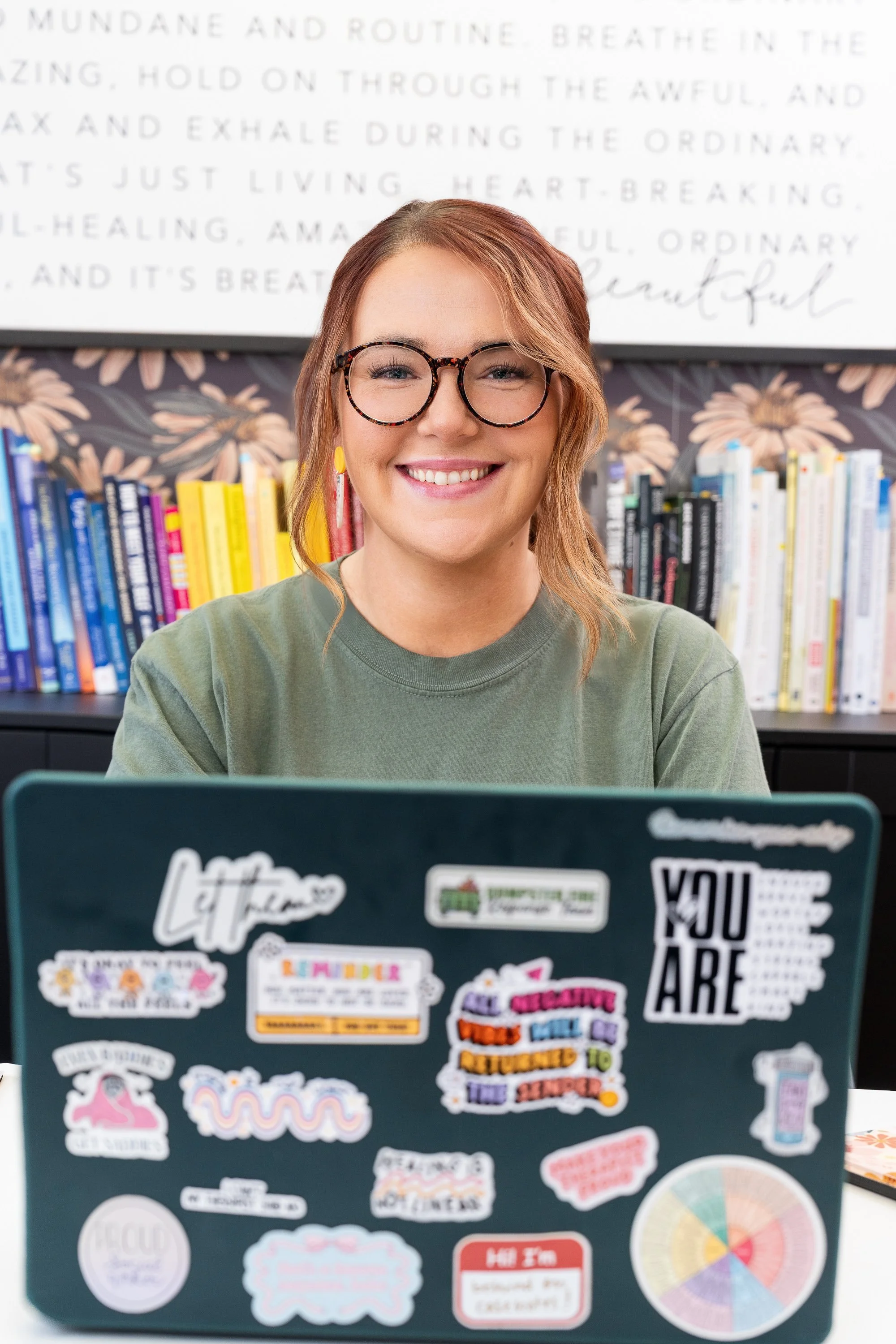 A woman with glasses and earrings sitting at a desk with a laptop covered in colorful stickers. She is smiling, wearing a green shirt, with a colorful bookshelf and decorative wallpaper in the background.