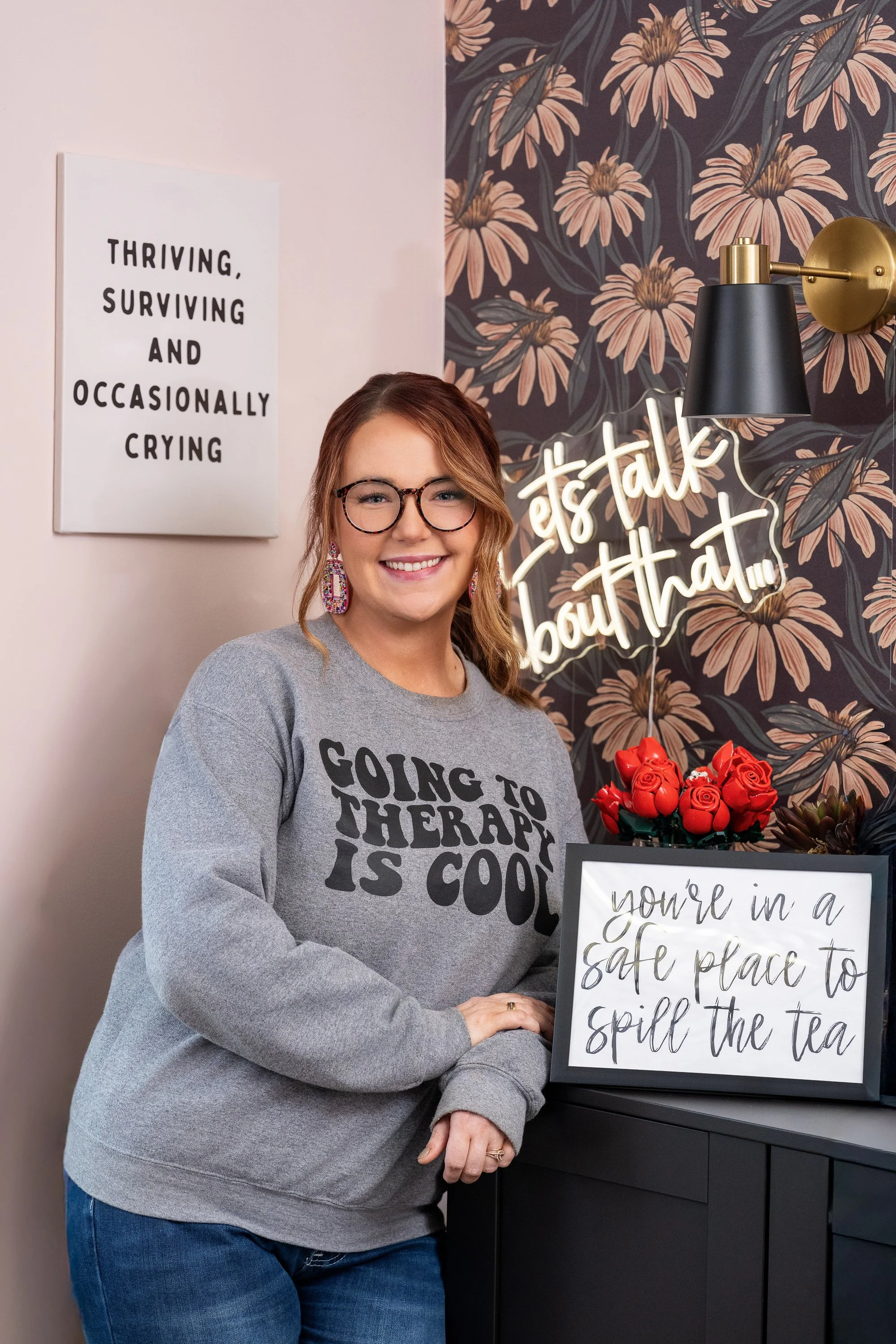 A woman with glasses and earrings, wearing a gray sweatshirt that says 'Going to therapy is cool,' is smiling while standing next to a black table with a bouquet of red roses and a framed sign that reads 'you're in a safe place to spill the tea.' Behind her is a floral wallpapered wall with a neon sign saying 'let's talk about that' and a gold and black wall sconce. To the left, there is a wall art with text 'Thriving, surviving and occasionally crying.'