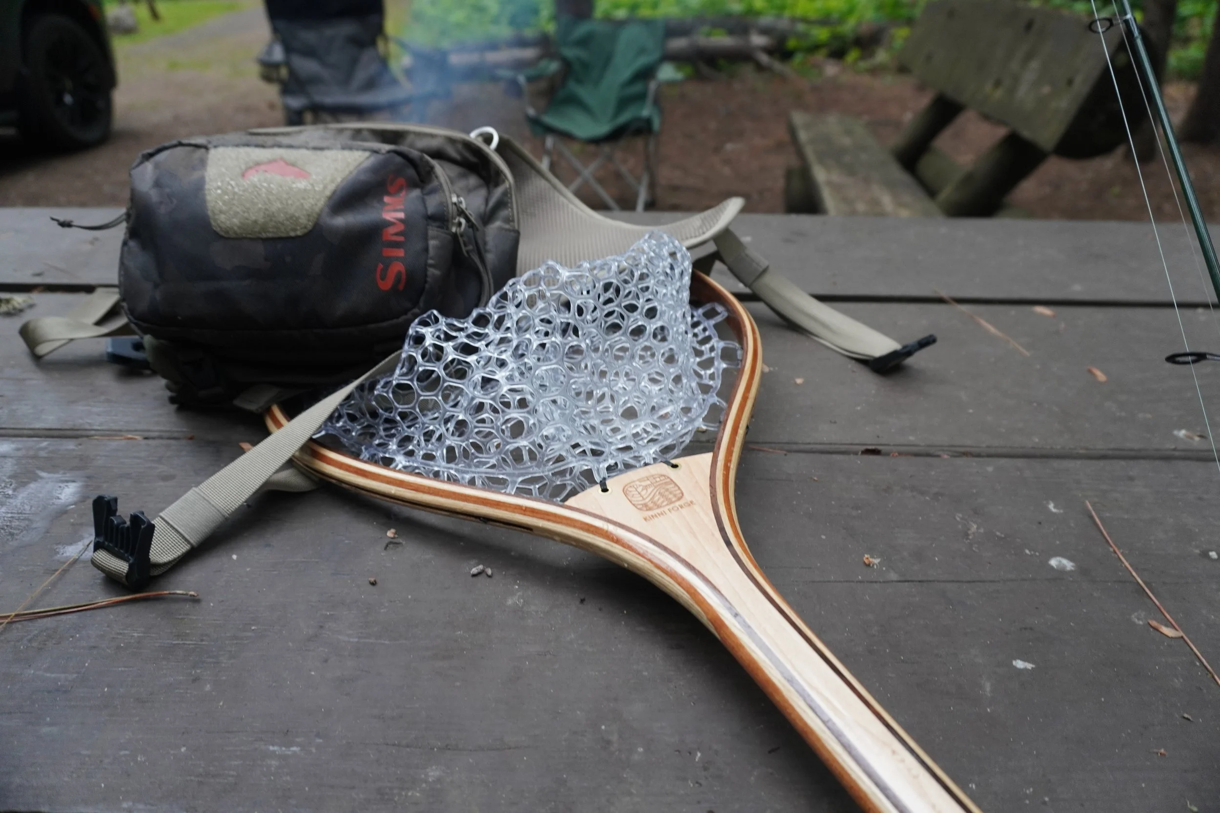 Hand crafted steam bent wood landing net for trout sitting on a camping table with fishing gear.