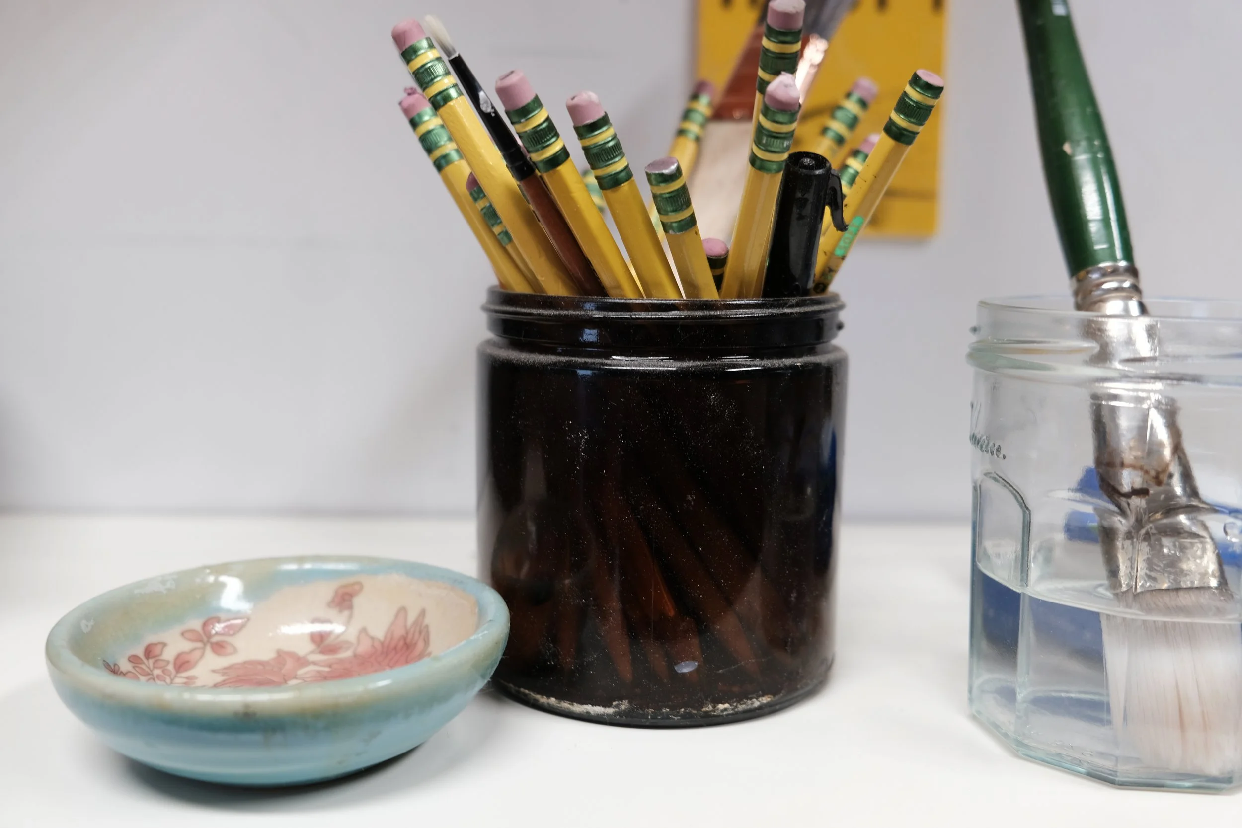 A black jar holding yellow pencils with erasers, black ballpoint pen, and a brown colored pencil, with a small decorative bowl beside it and a glass jar holding brushes and painting tools.