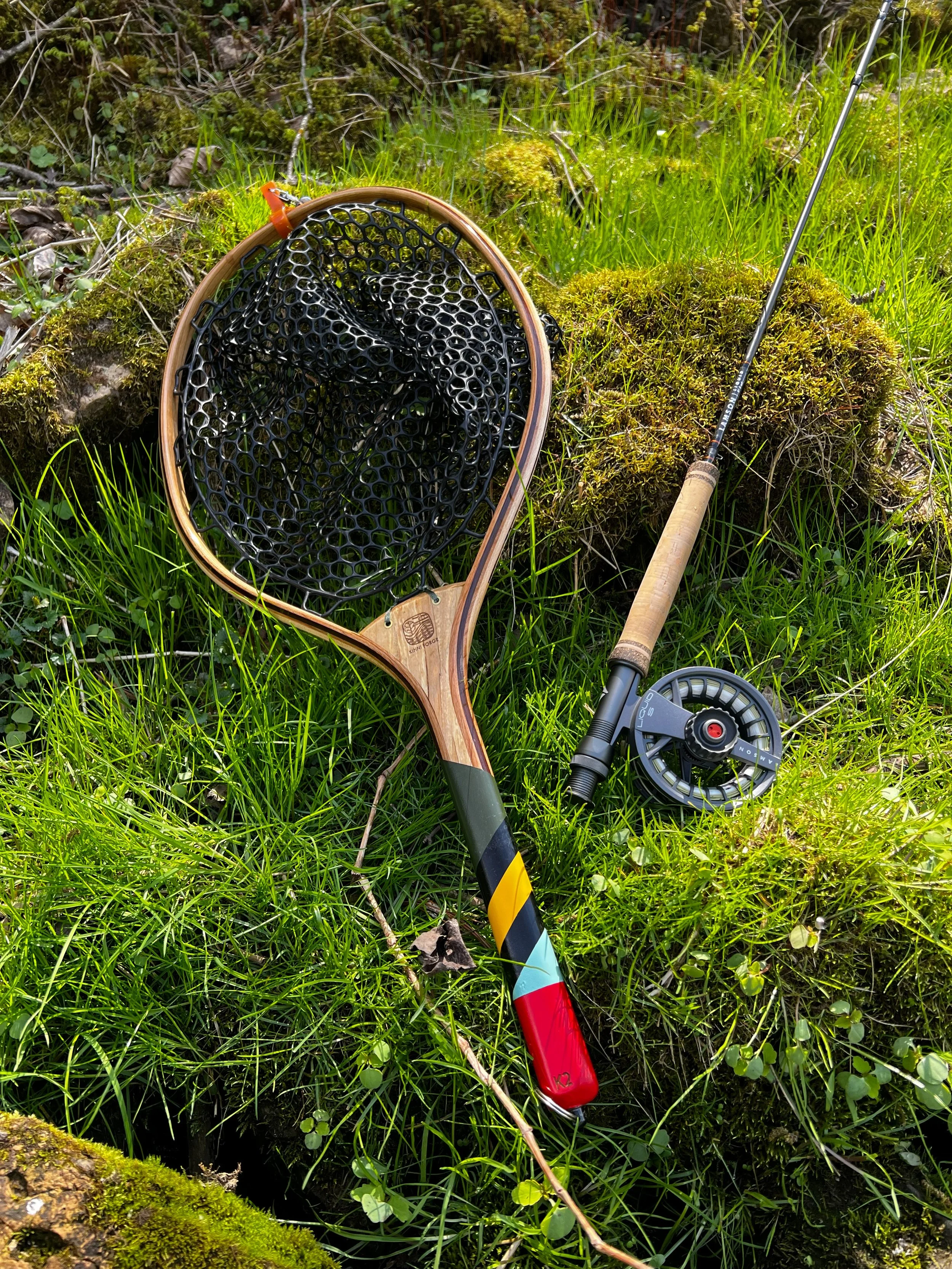 A fishing net, a fishing rod, and a reel lying on green grass and moss in a forested area.