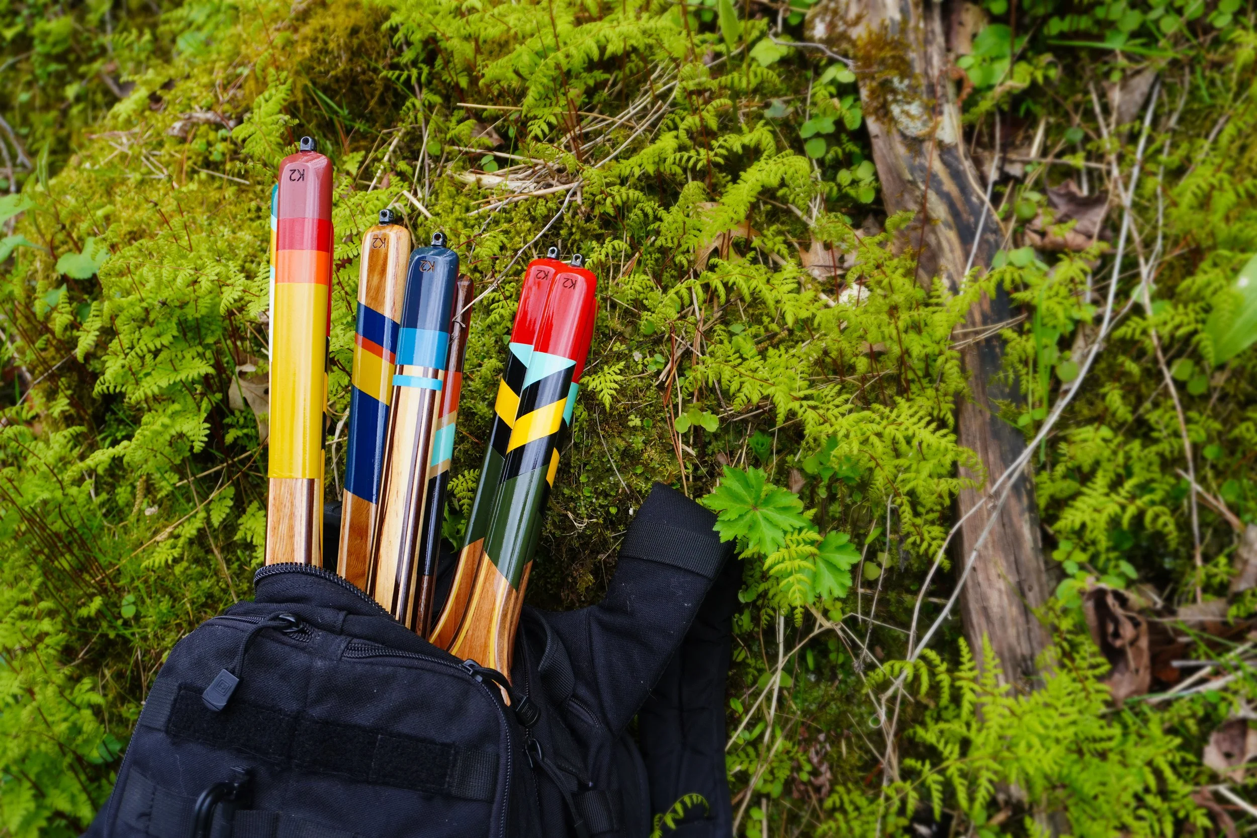A person wearing black pants and a black backpack standing on green ferns and mossy ground, holding five colorful hiking poles with wood, blue, red, and yellow designs.