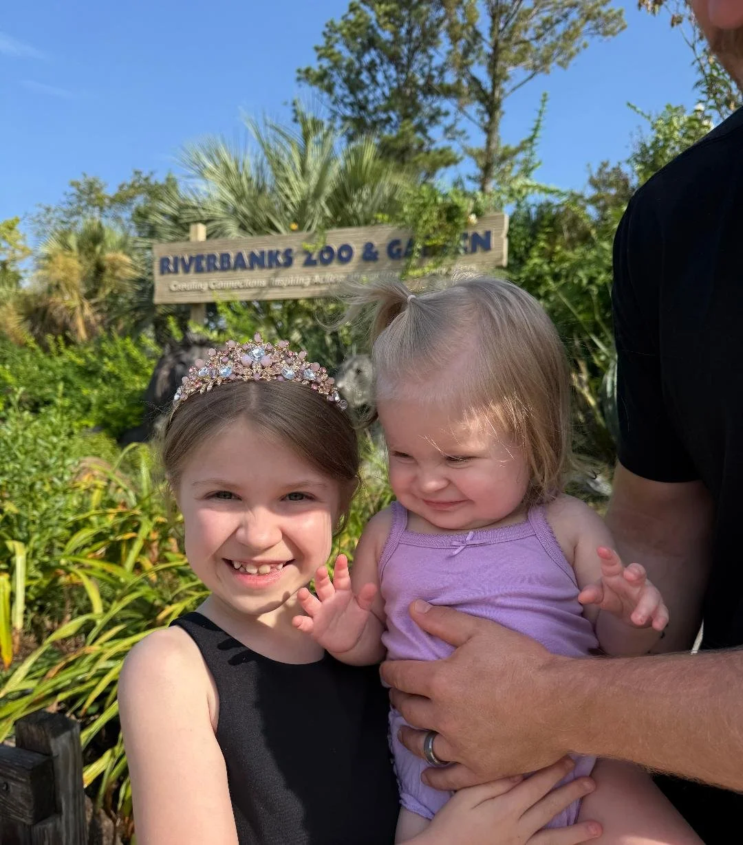 Two young girls, one wearing a tiara and a black dress, smiling at the camera, at Riverbanks Zoo & Garden.