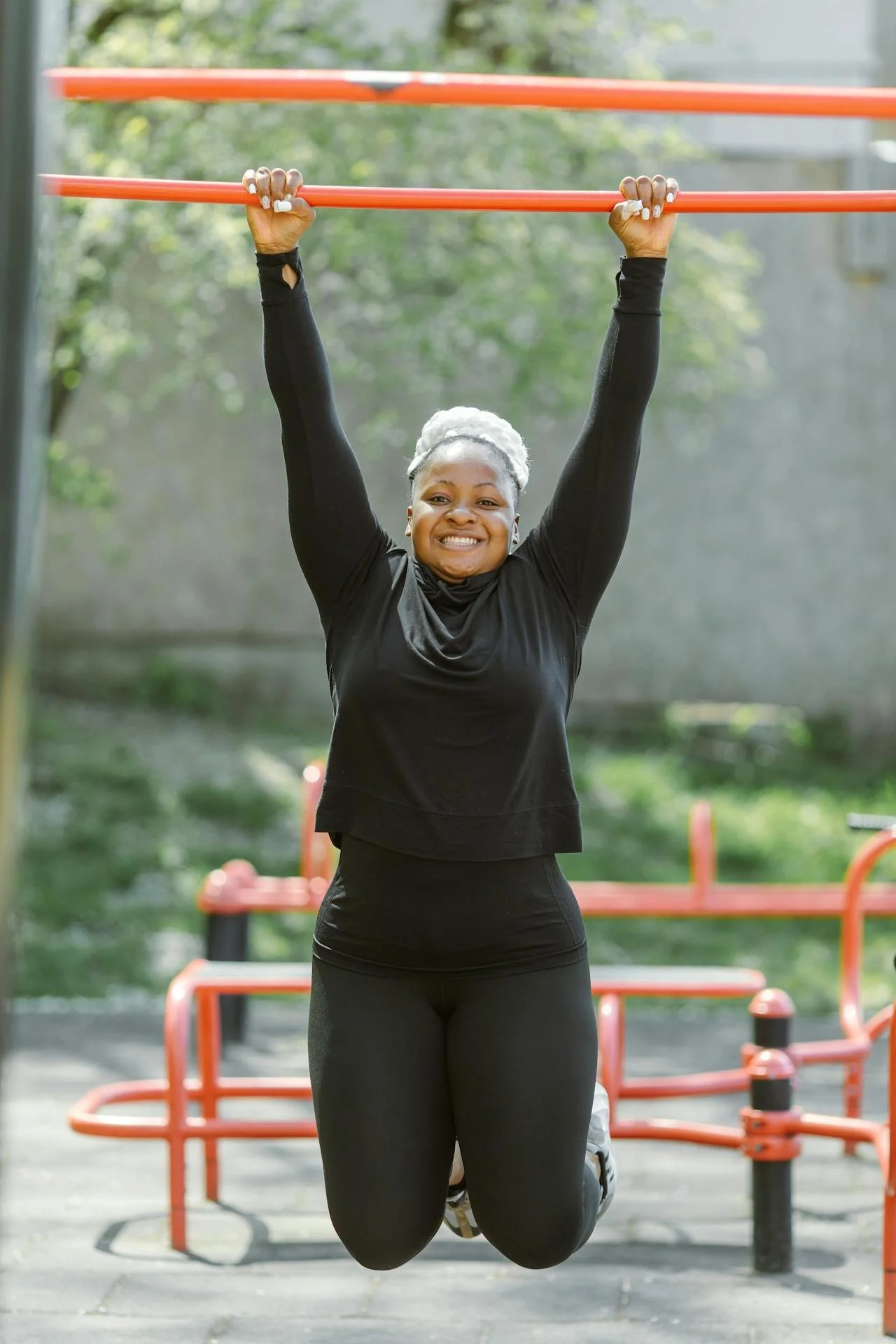 Black woman, about 35 years old, hanging from monkey bars in a park, smiling and enjoying physical activity.