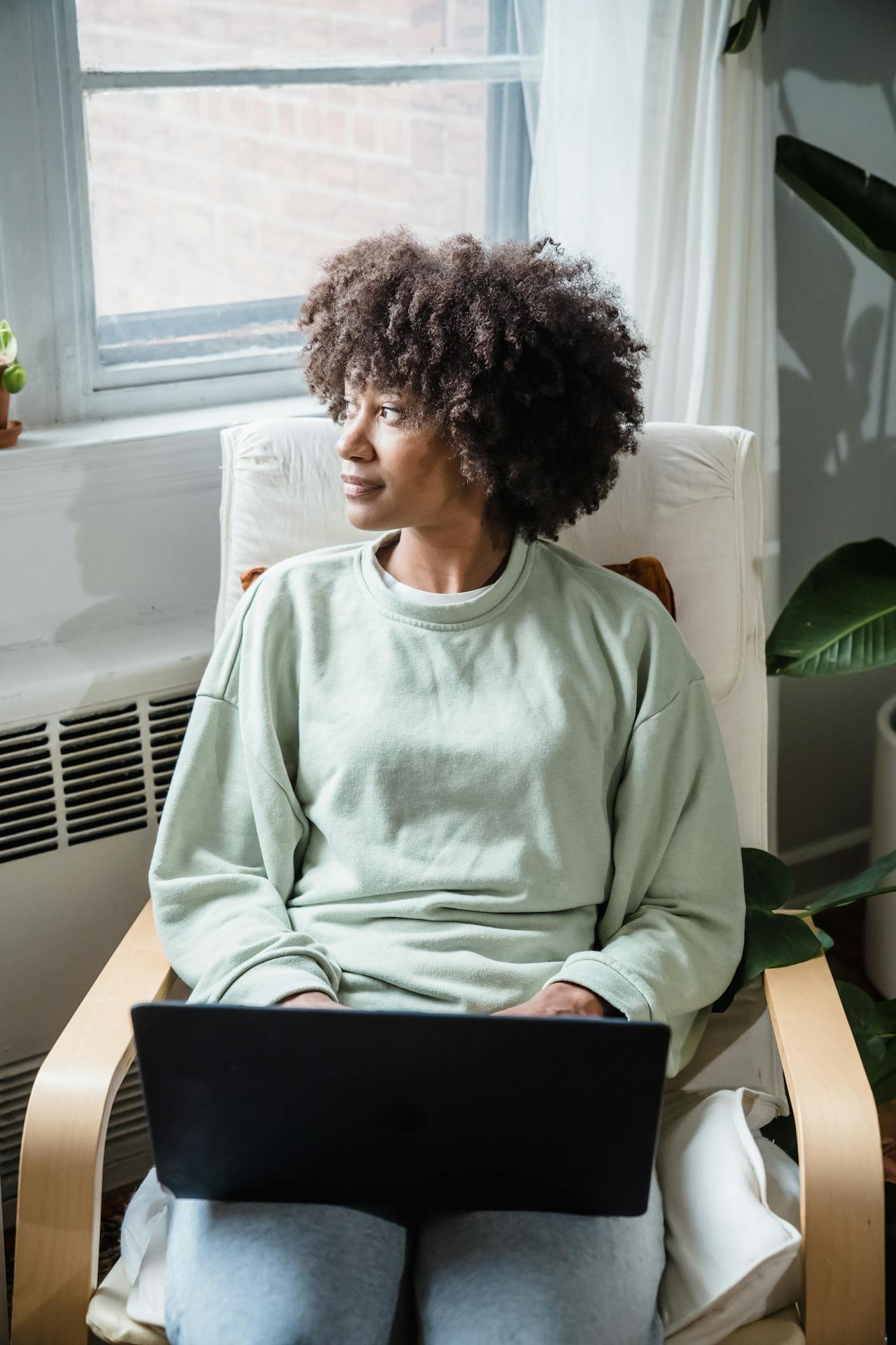 Black woman, about 35 years old, staring thoughtfully out of a window with a laptop on her lap, hands resting on the laptop, appearing contemplative.