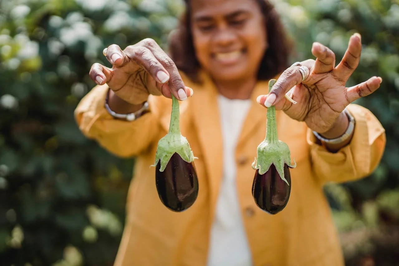 Middle-aged Black woman smiling and holding two eggplants by their stems, symbolizing ovaries.