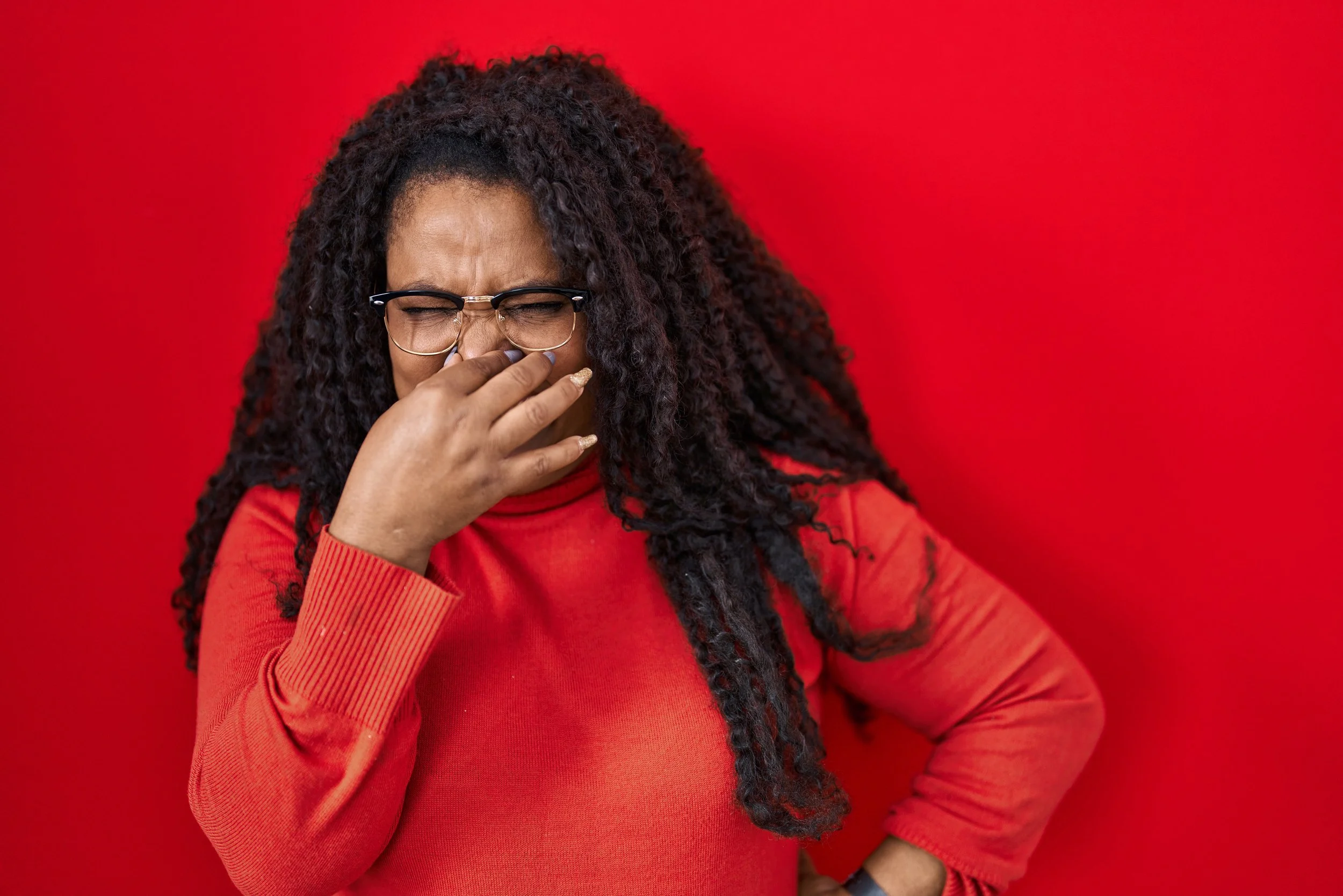 Black woman with locs wearing a red shirt and holding her nose, standing against a plain red background.