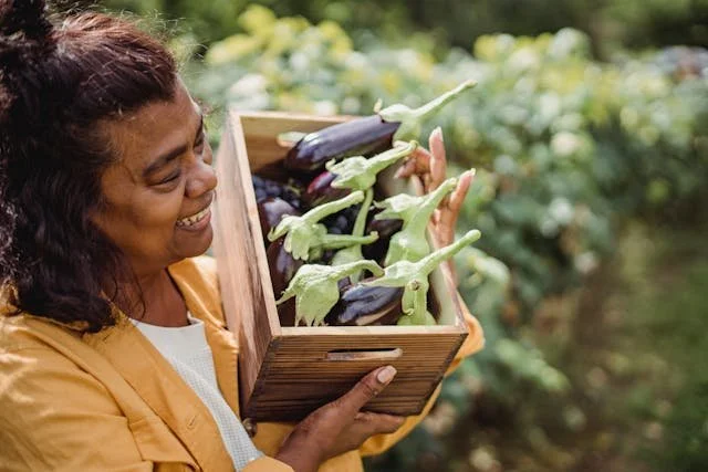 Black woman smiling while holding a wooden box with eggplants inside, symbolizing growth and vitality.