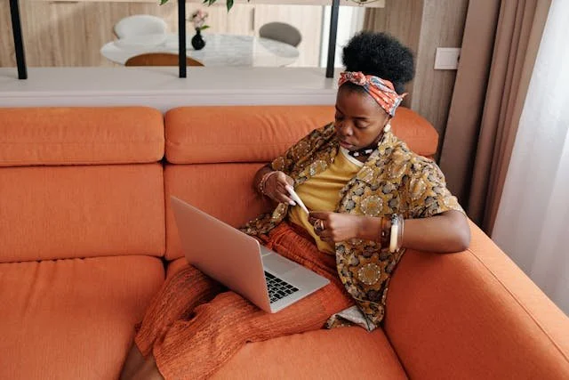 Black woman sitting on a couch holding a paper in one hand and a laptop in the other, looking focused.