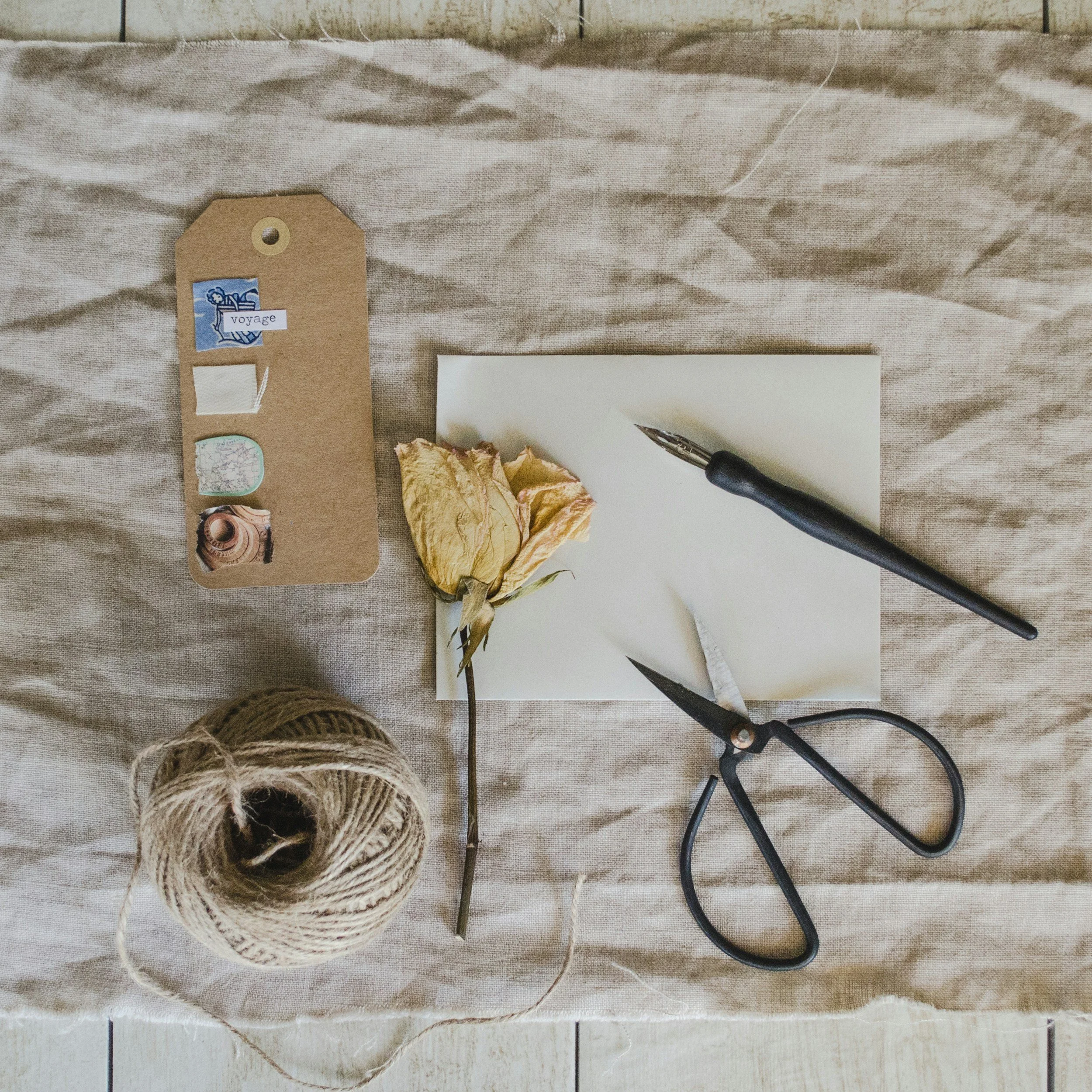Dried yellow flower, pair of scissors, fountain pen, blank notepad, roll of twine, paper tag with travel-themed stickers on textured beige fabric.