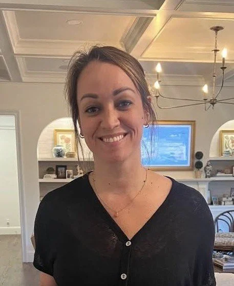 A woman with brown hair tied back, wearing a black top, smiling inside a home with a chandelier and a window showing a blue sky.